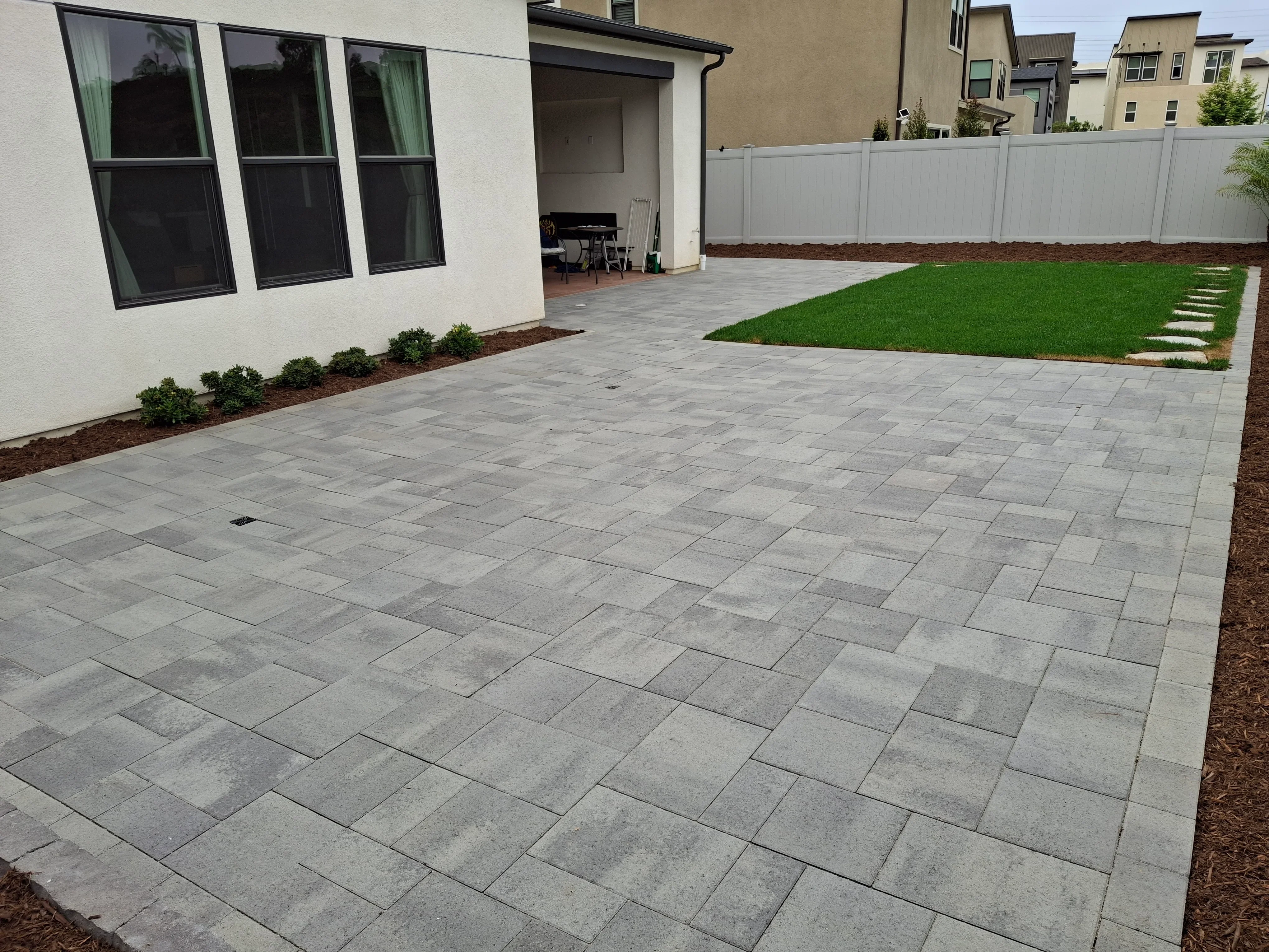 Gray concrete paver patio with running bond pattern, sod lawn, and white vinyl fence