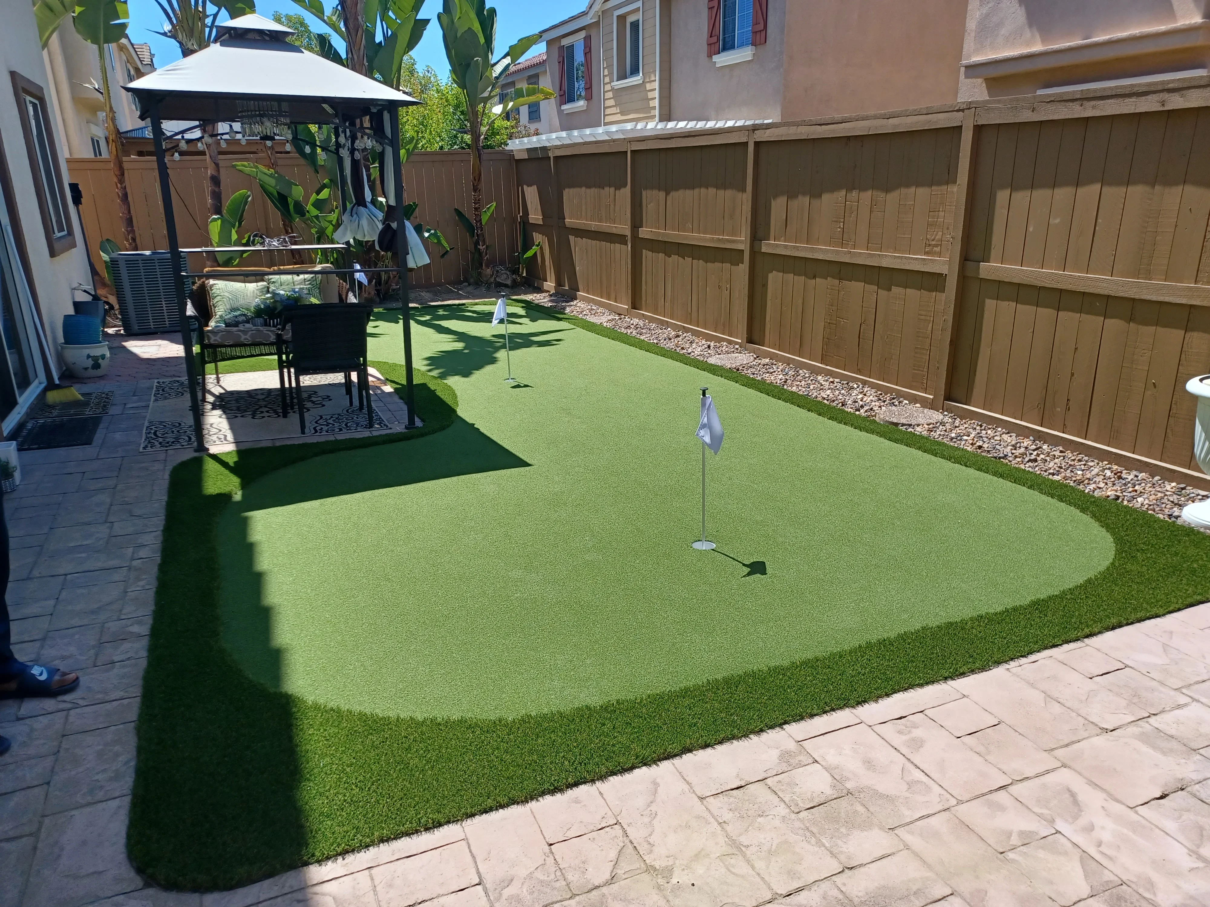 Synthetic turf putting green with two holes, flagsticks, river rock border, and wood fence