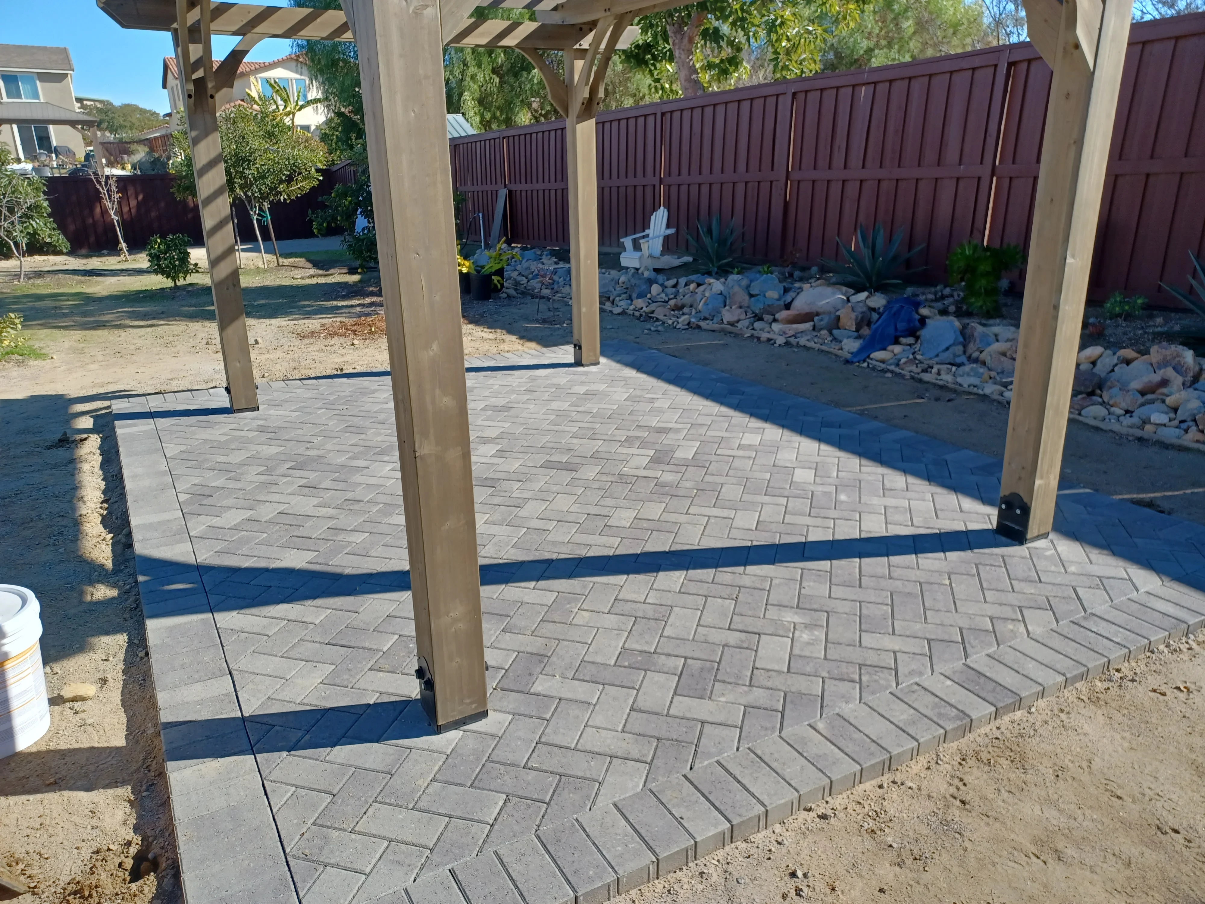 Wooden pergola over gray permeable pavers in herringbone pattern with rock landscaping and metal fence