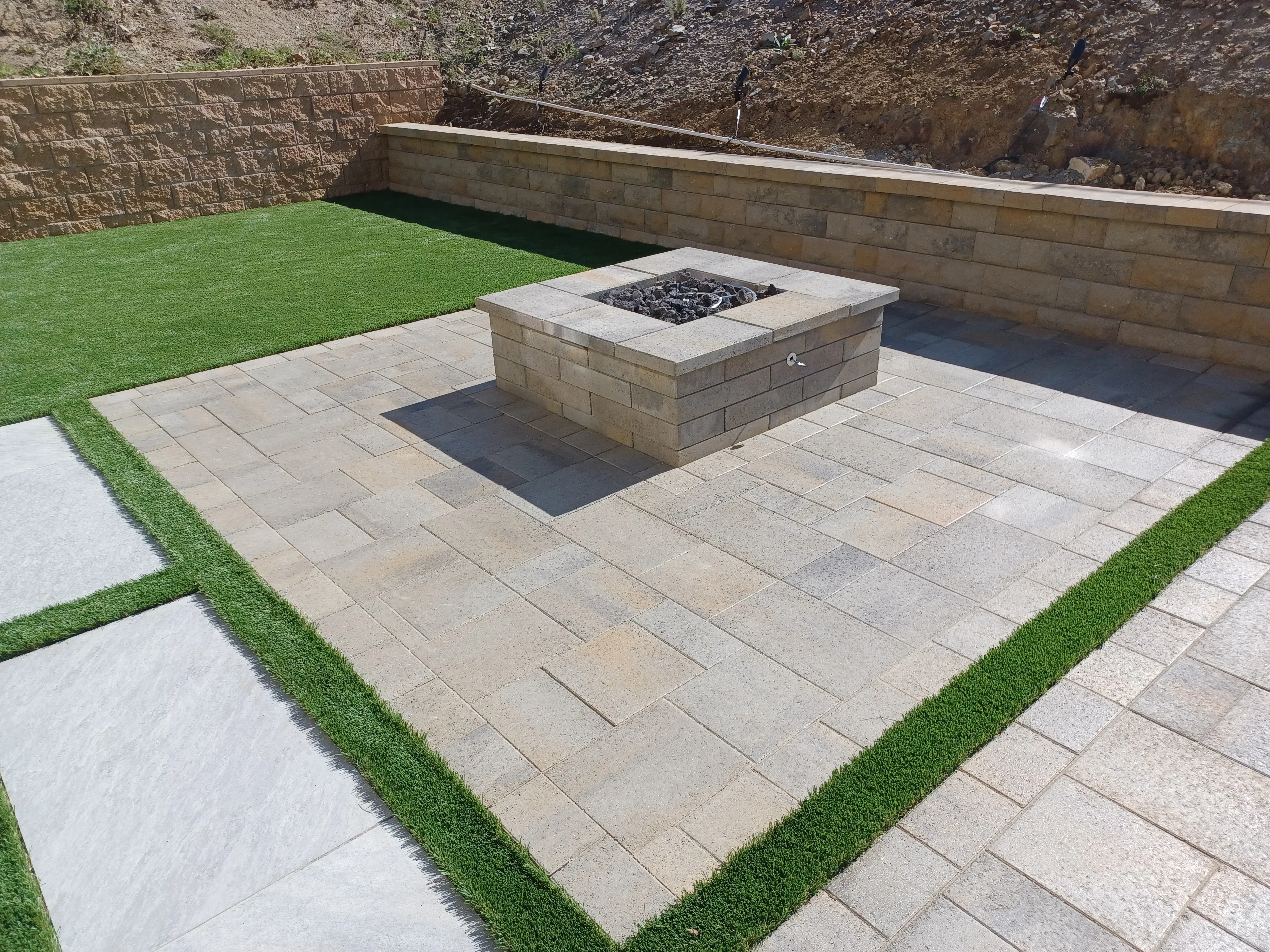 Stone paver patio with built-in fire pit, retaining walls, and artificial turf borders