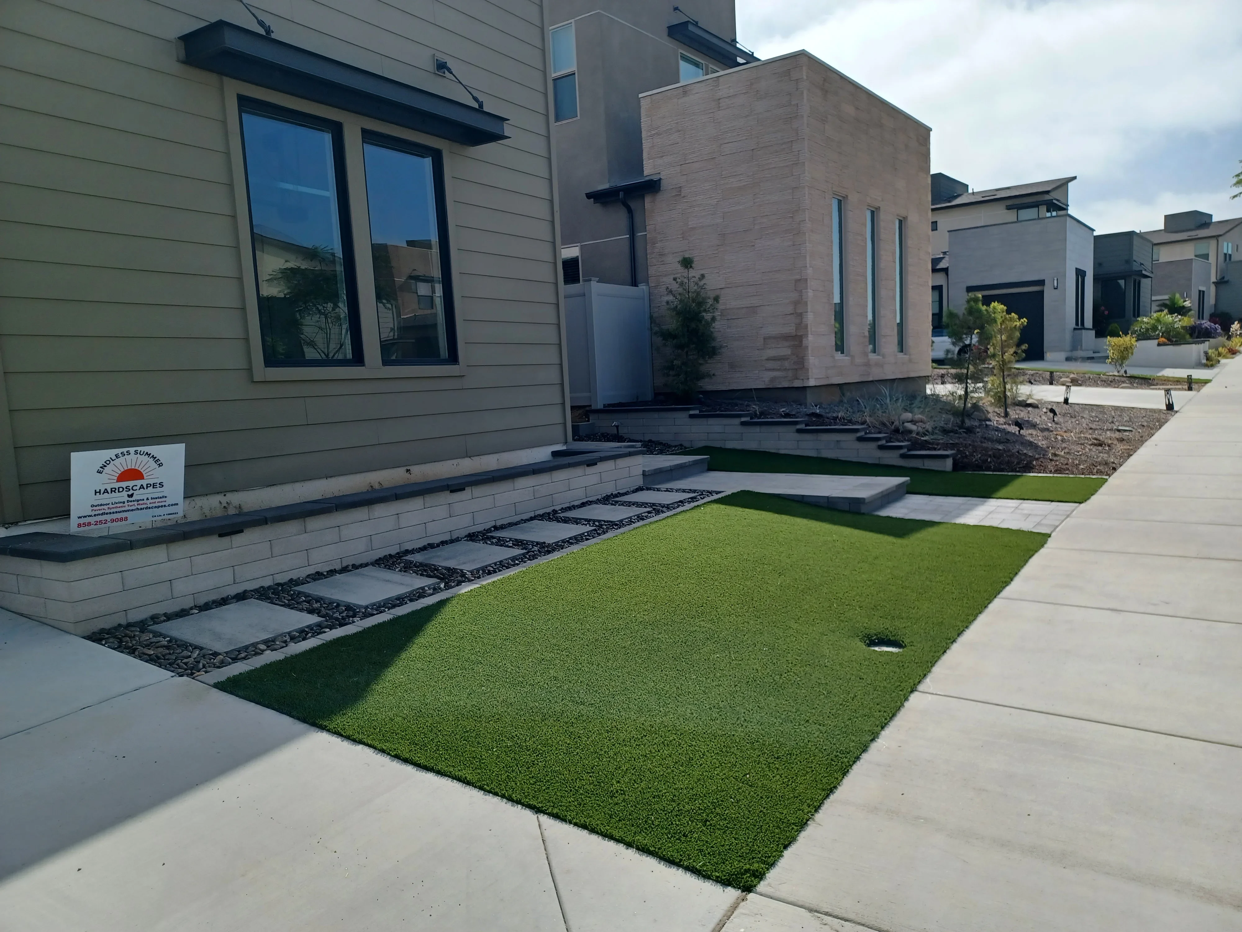 Concrete paver walkway with black gravel, synthetic turf lawn, and planter beds at modern home