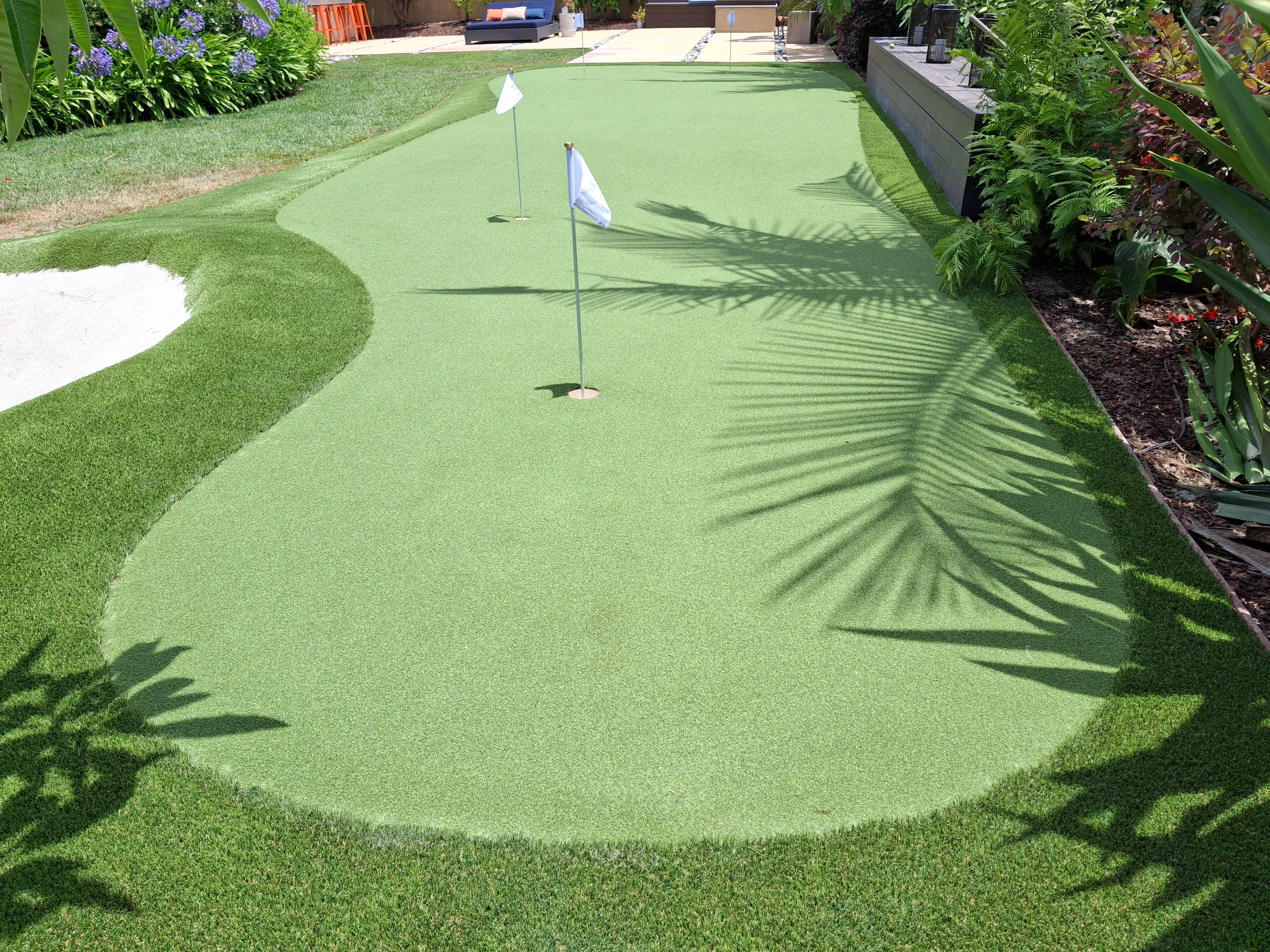 Synthetic putting green with sand bunker, two flagged holes, and natural turf borders in residential backyard