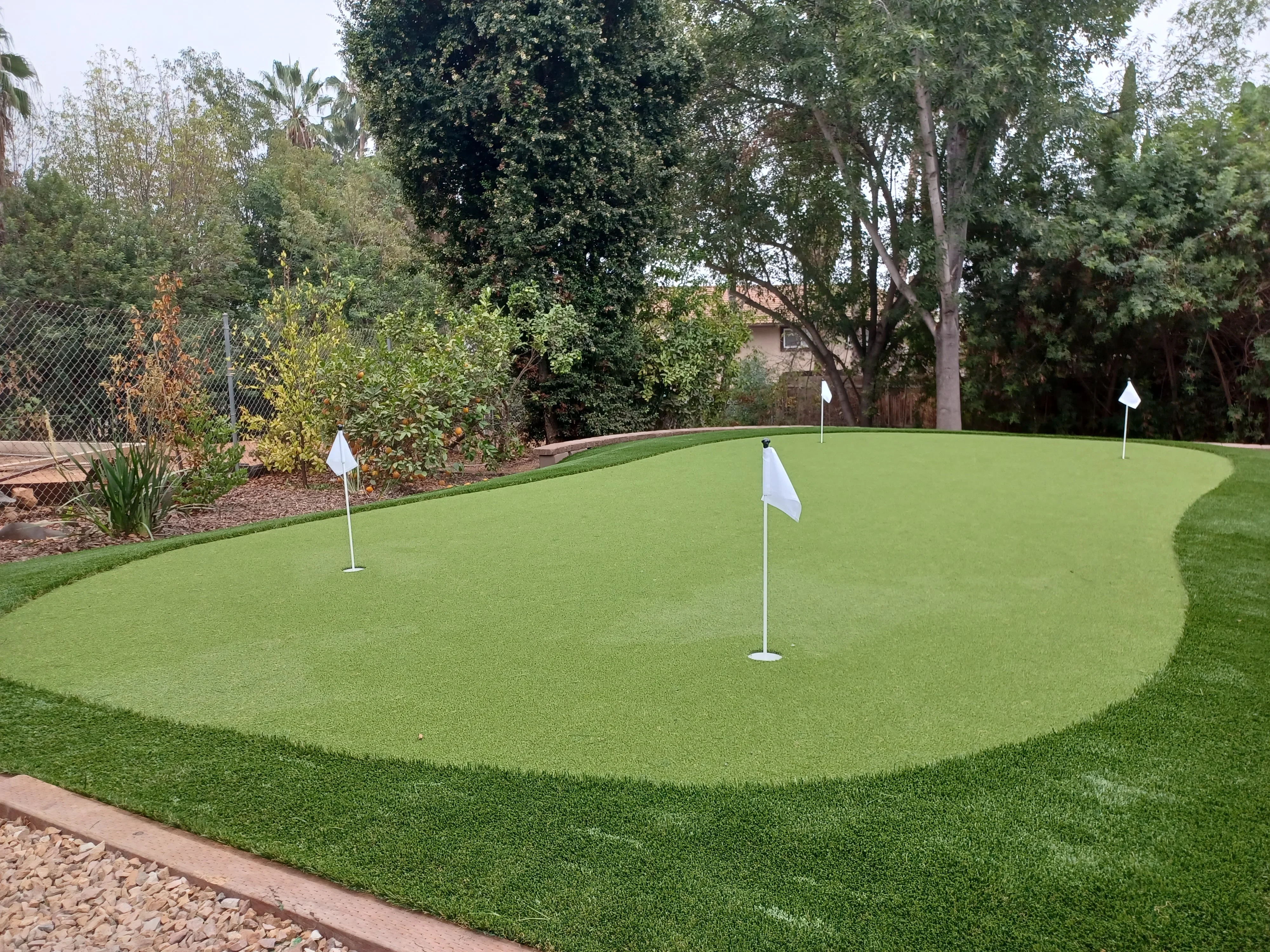 Synthetic turf putting green with flagsticks, rock edging, and mature trees
