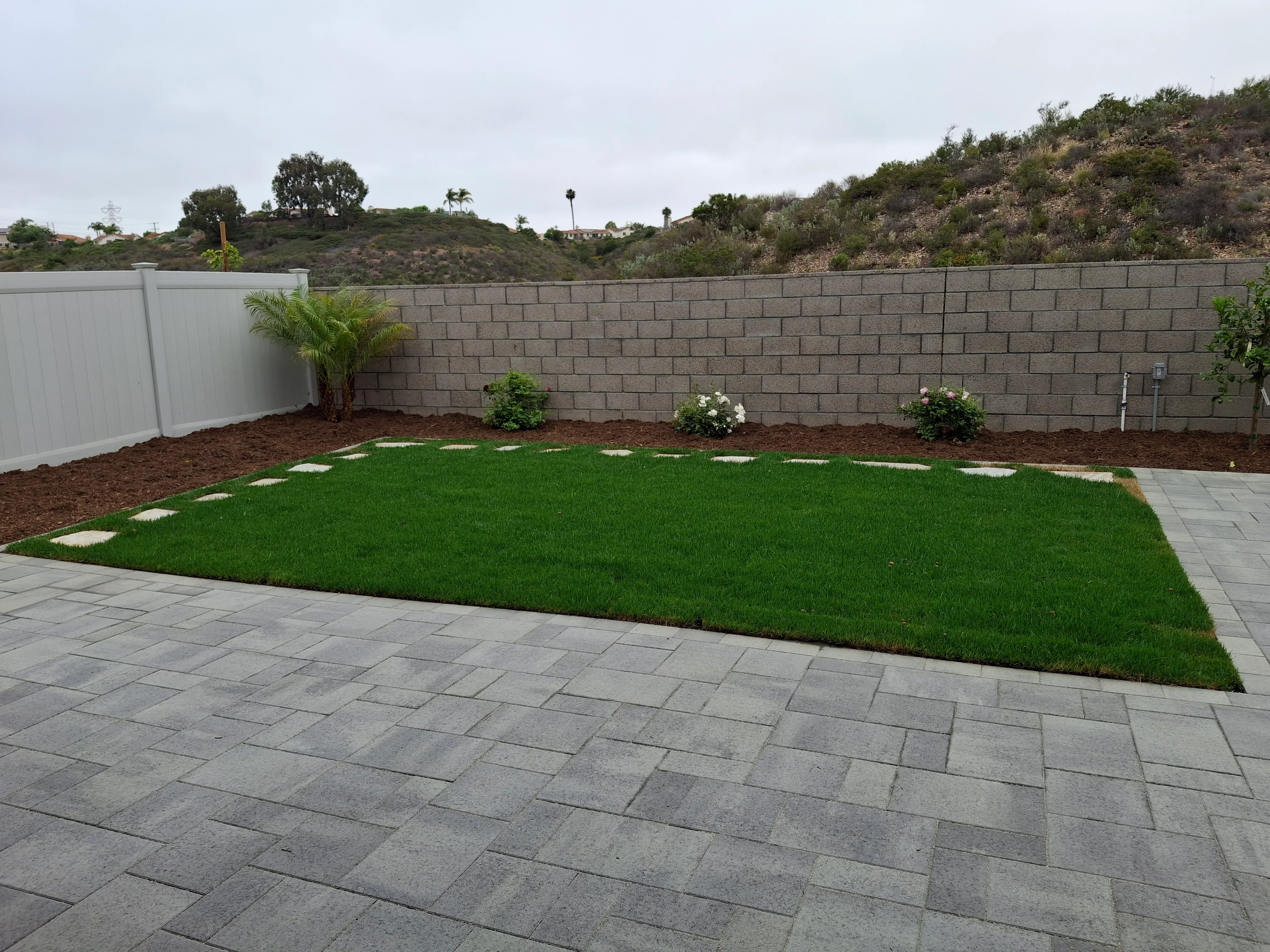 Gray concrete paver patio, new lawn with stepping stones, concrete block retaining wall, and mulch beds