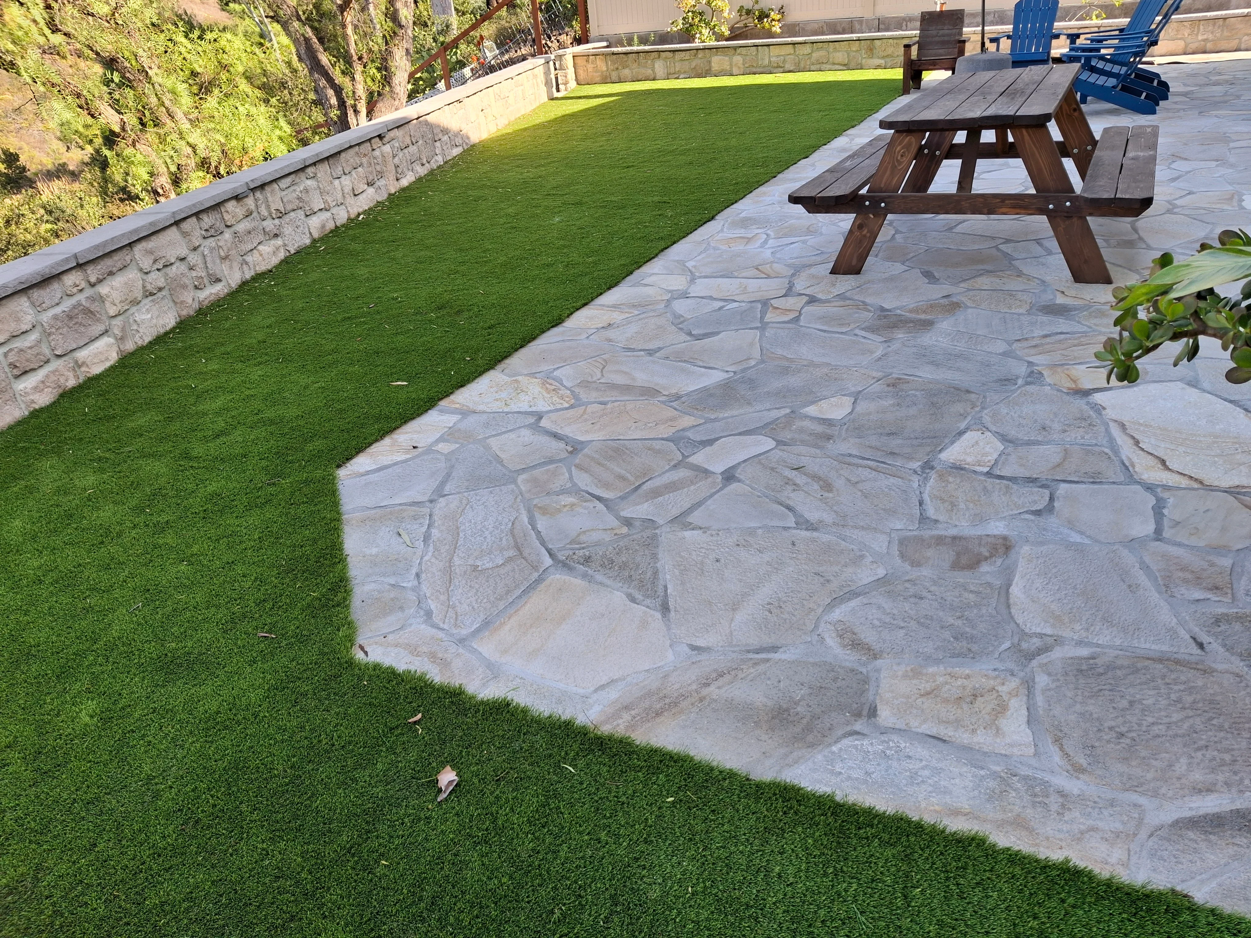 Flagstone patio with irregular stone pattern, synthetic turf lawn, and stone retaining wall with wooden picnic table