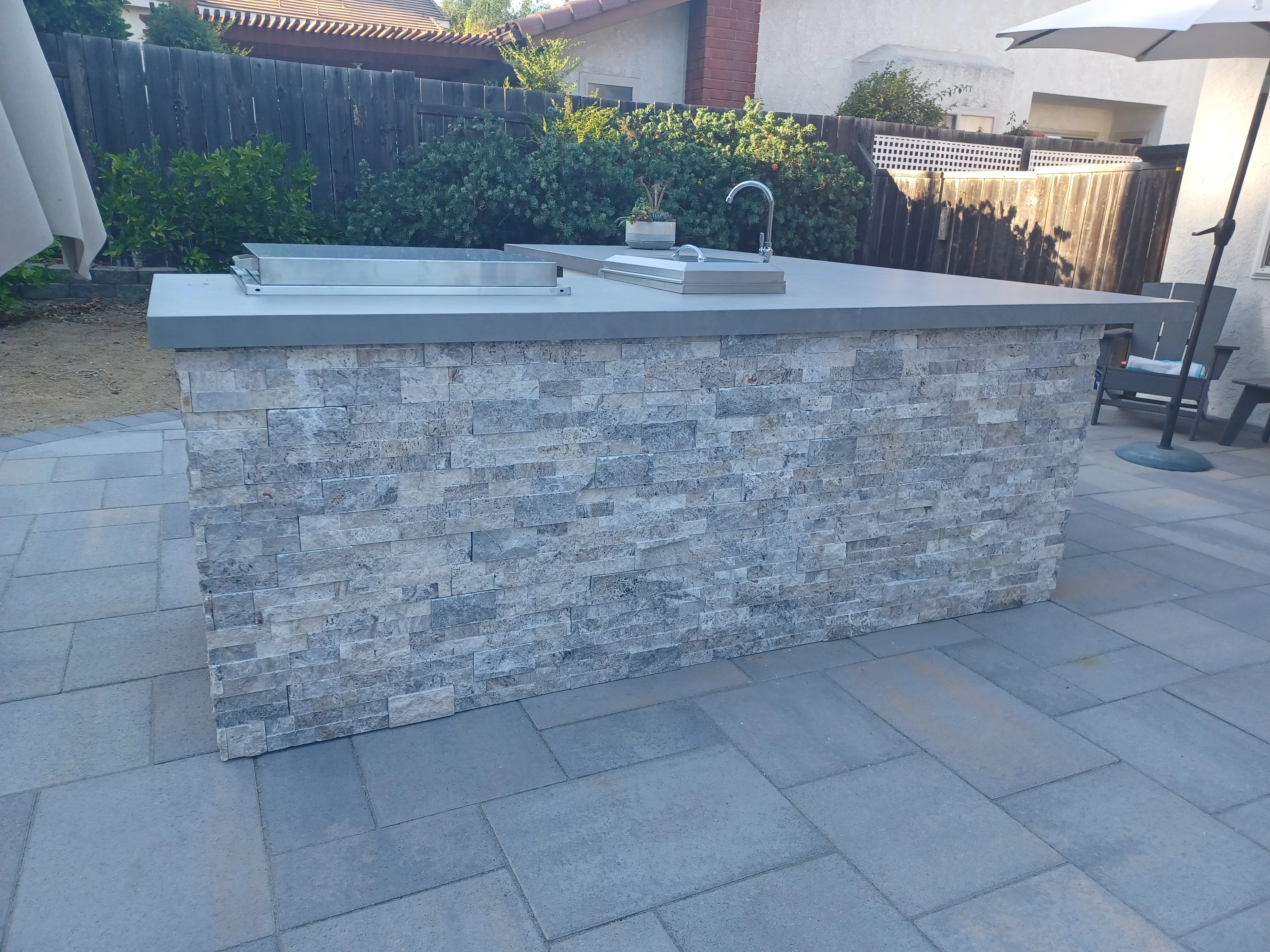 Stone veneer outdoor kitchen island with concrete countertop, sink, and faucet on paver patio