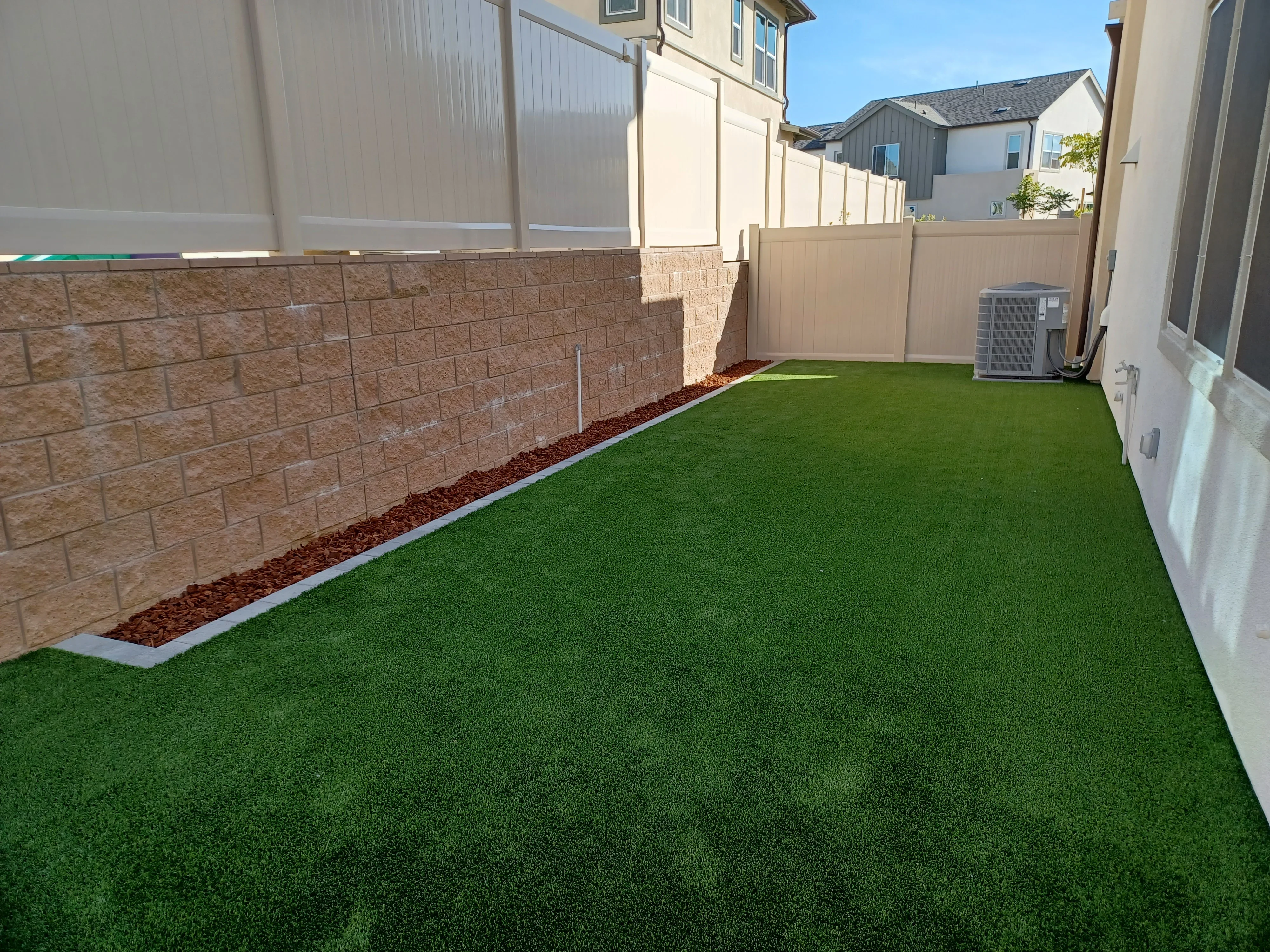 Synthetic turf lawn with stone retaining wall, red mulch borders, and vinyl fencing in residential backyard