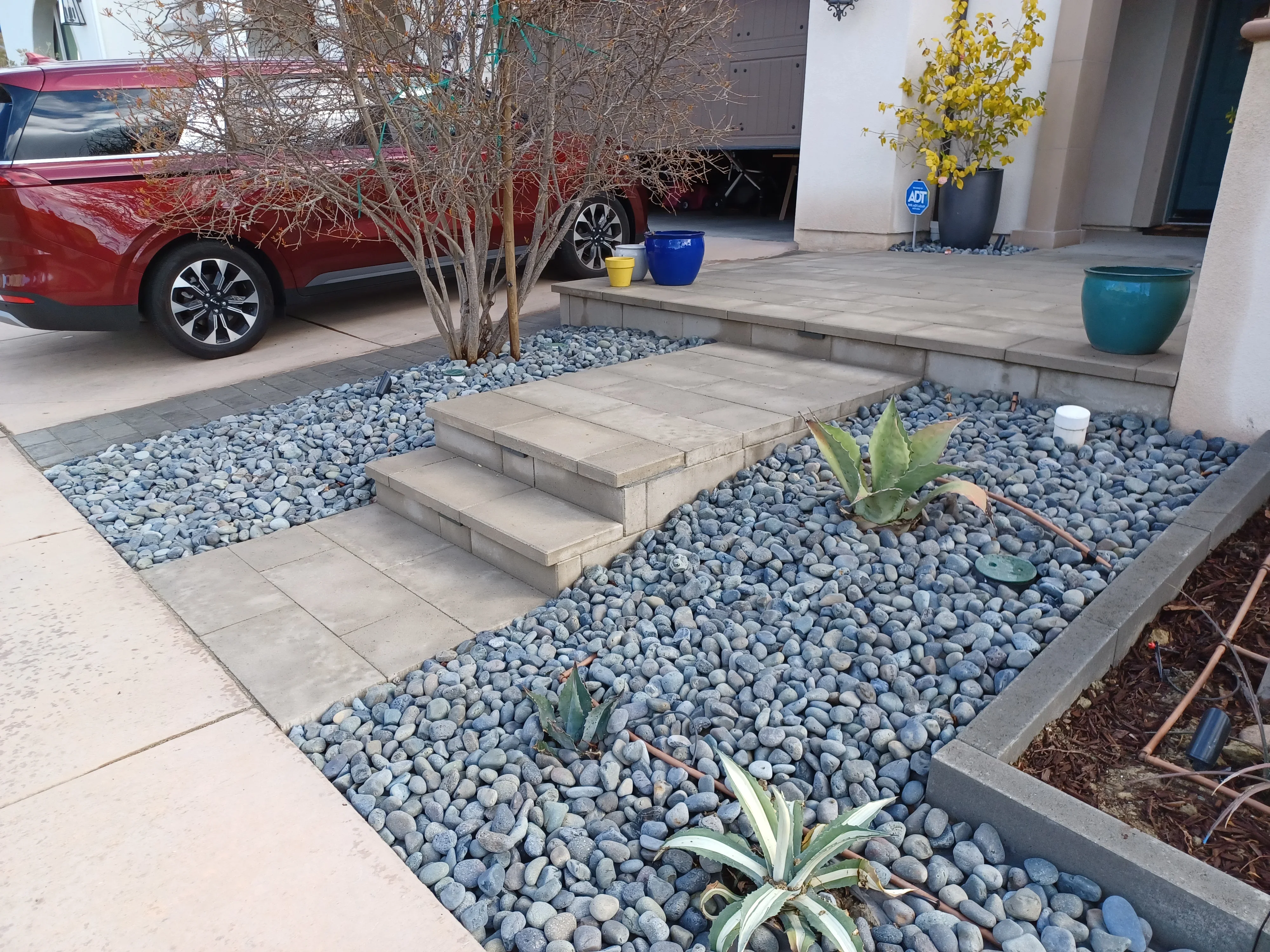 Paver driveway and patio with blue-gray river rock, steel-edged planting beds, and concrete steps
