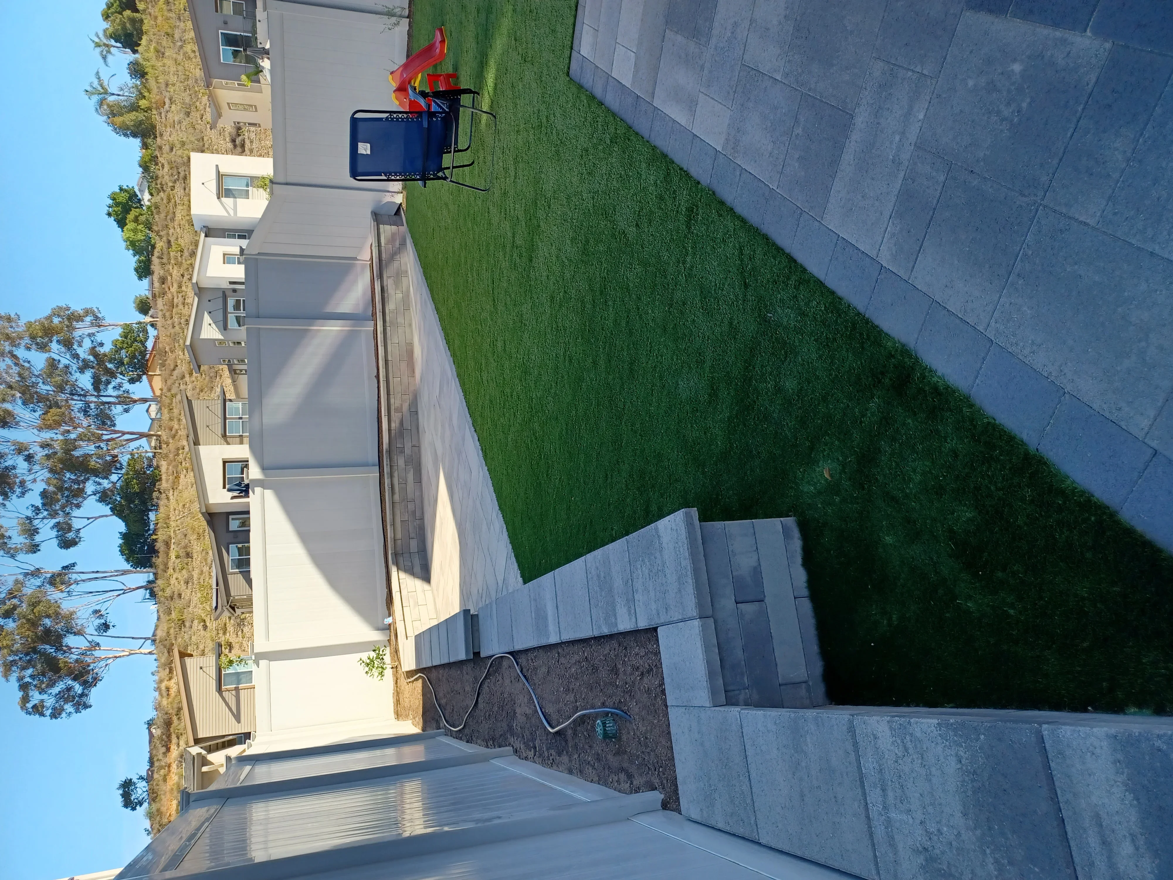 Gray paver patio with retaining wall, landscape fabric, and manicured lawn area adjacent to residential home