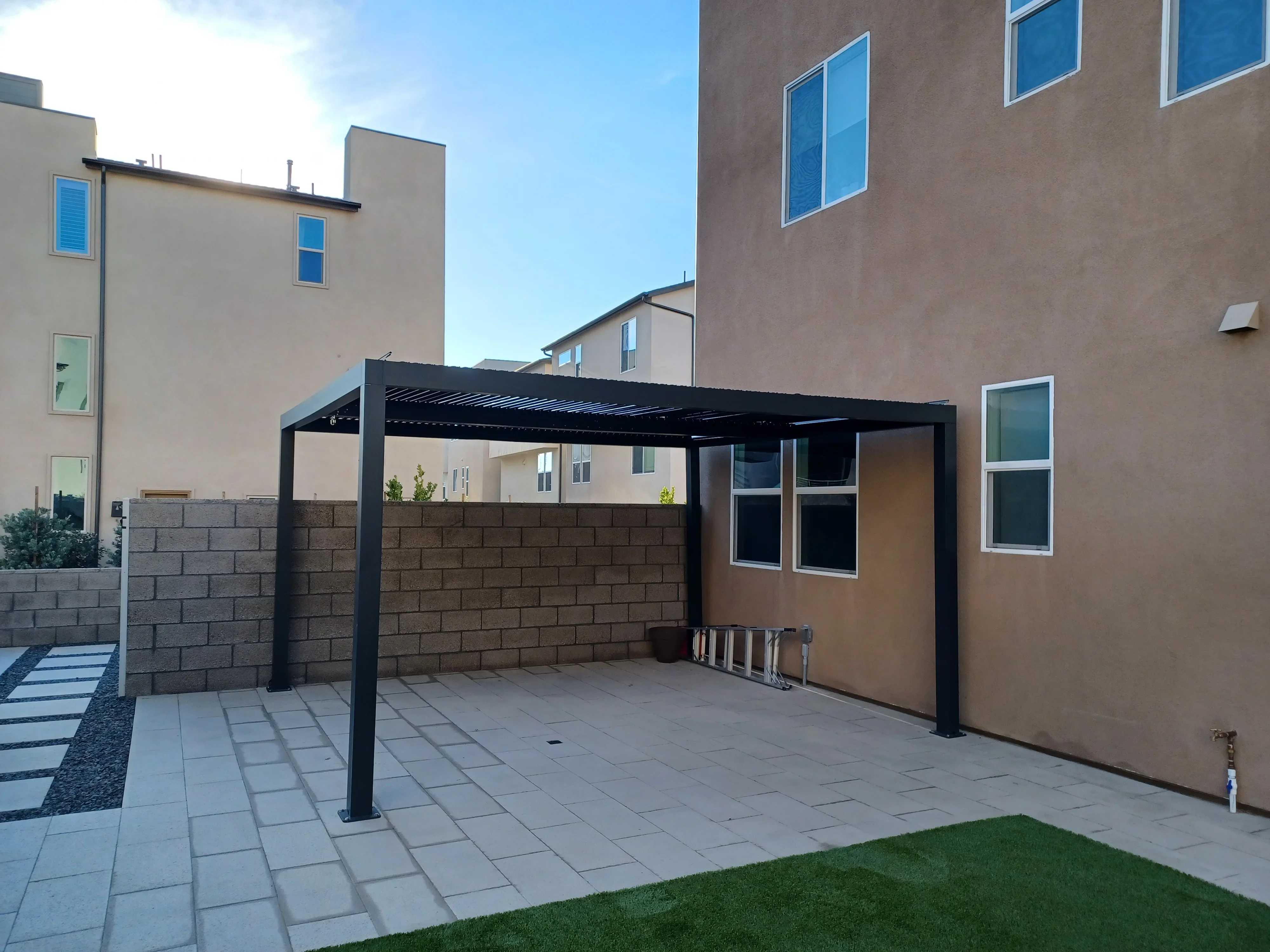 Black pergola with louvered roof over paver patio and concrete block retaining wall