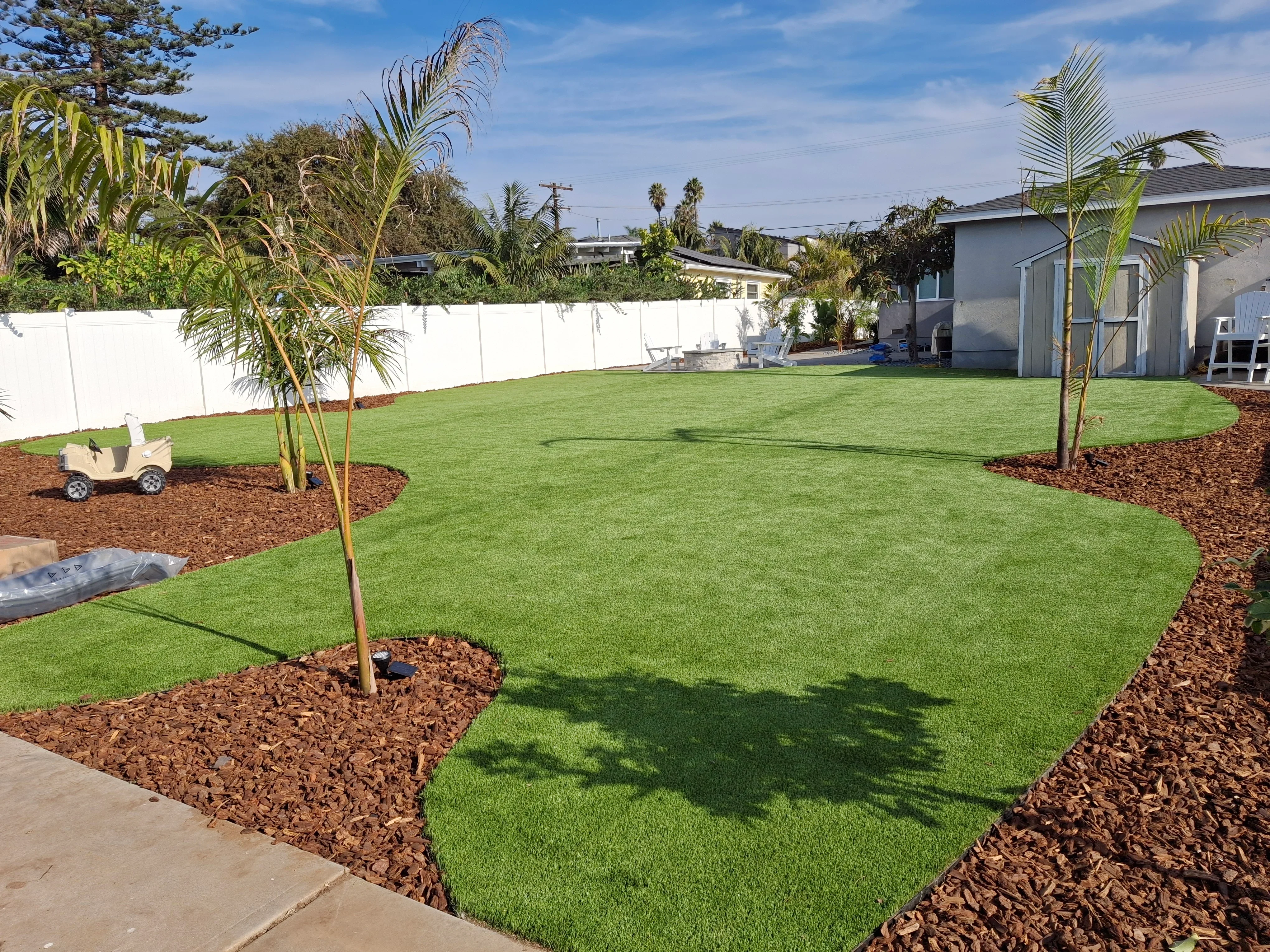 Synthetic turf lawn with mulch beds, palm trees, white vinyl fence, and concrete patio edging