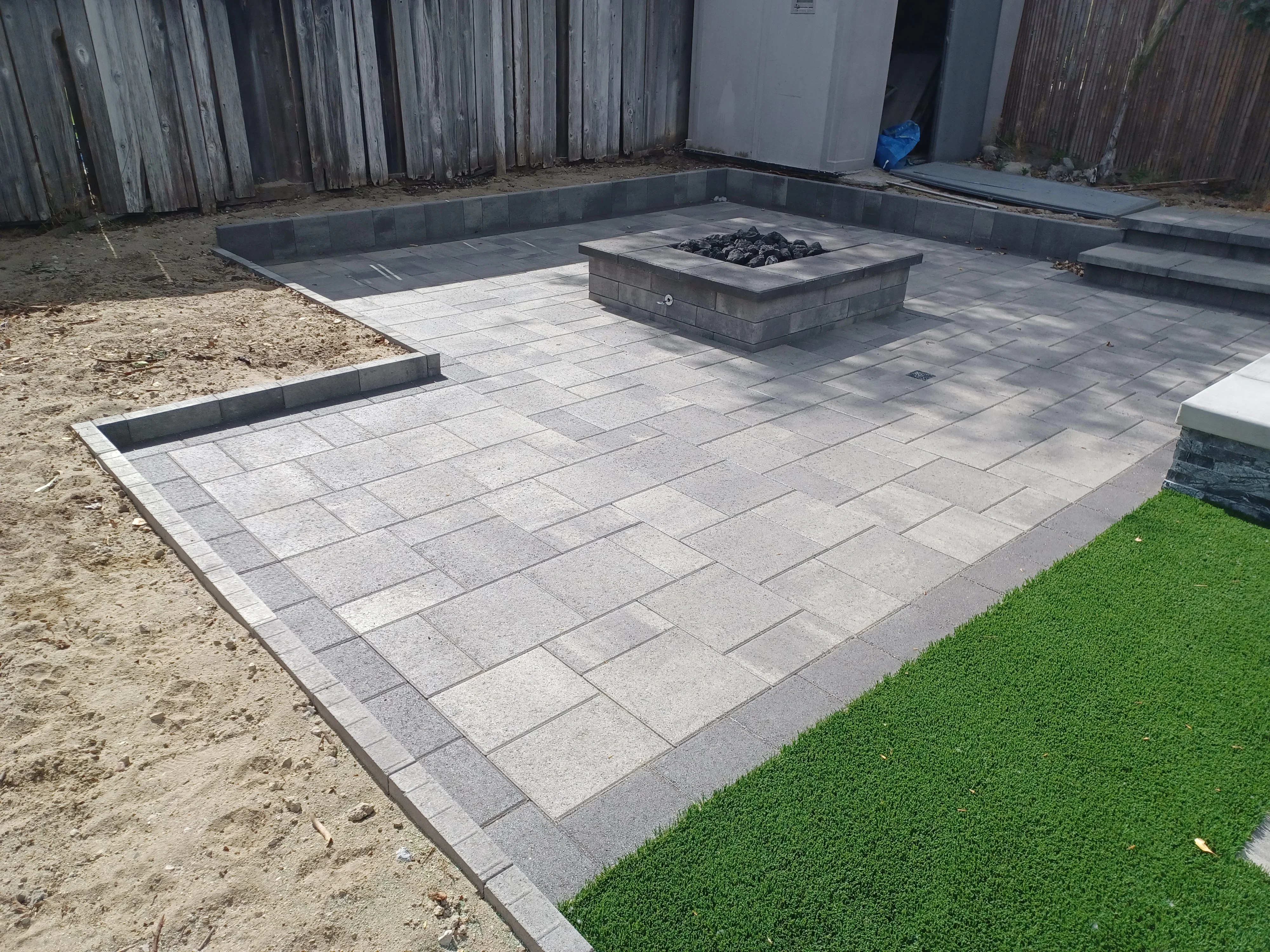 Gray concrete paver patio with dark border and central stone fire pit installation