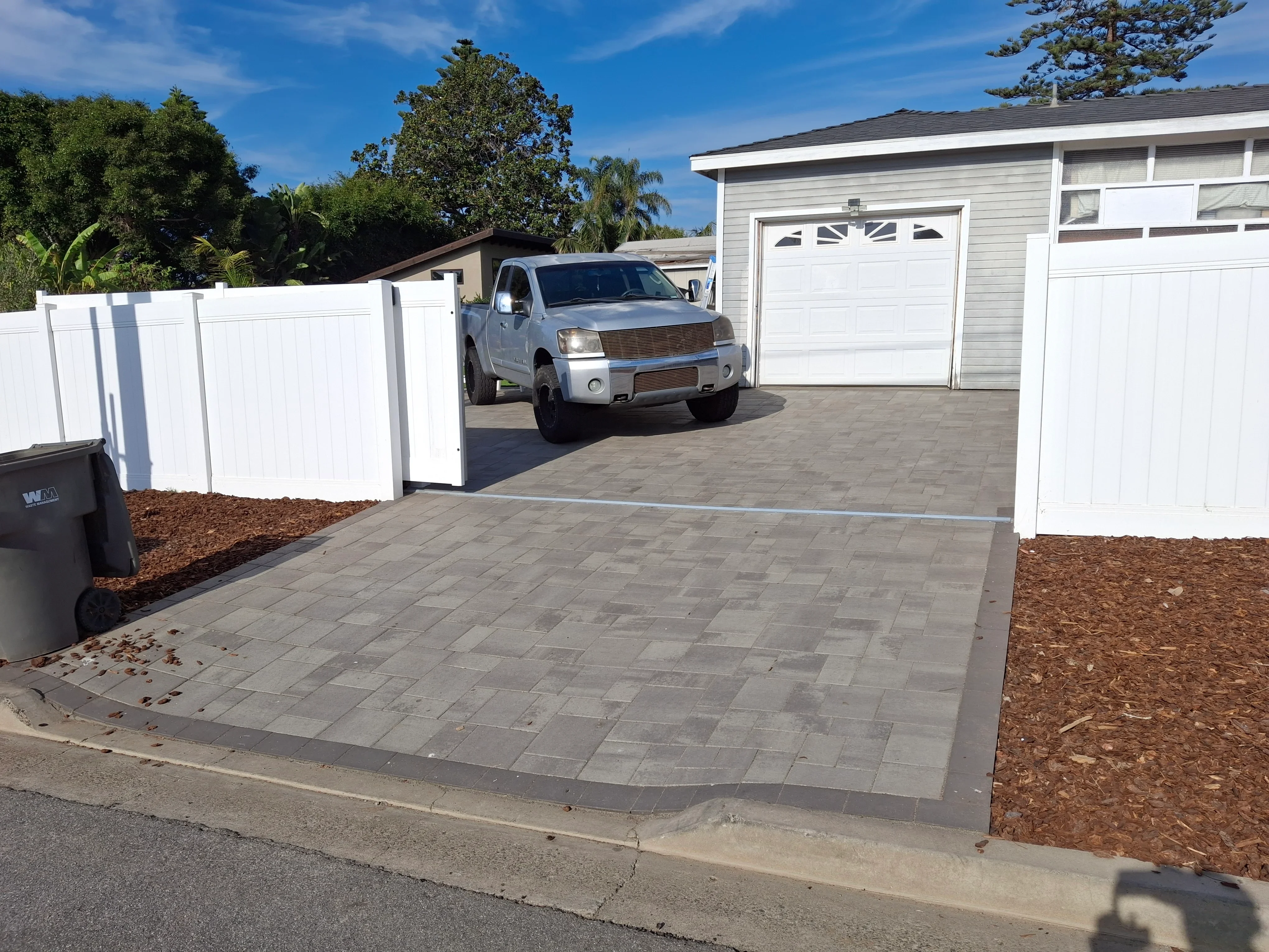 Gray permeable pavers driveway with white vinyl privacy fence and mulched landscaping