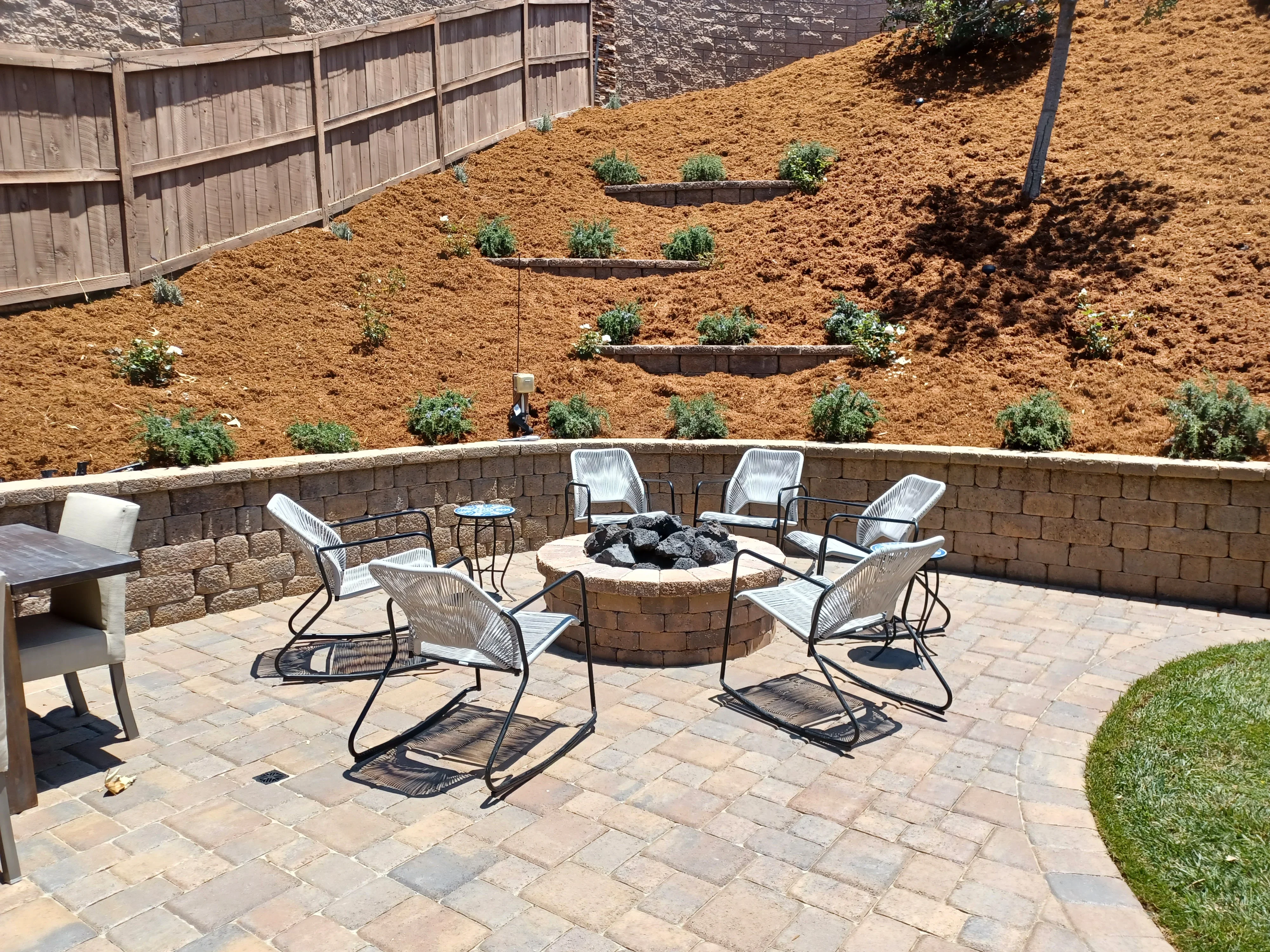 Paver patio with circular fire pit, stacked stone retaining wall, and tiered landscape beds on slope