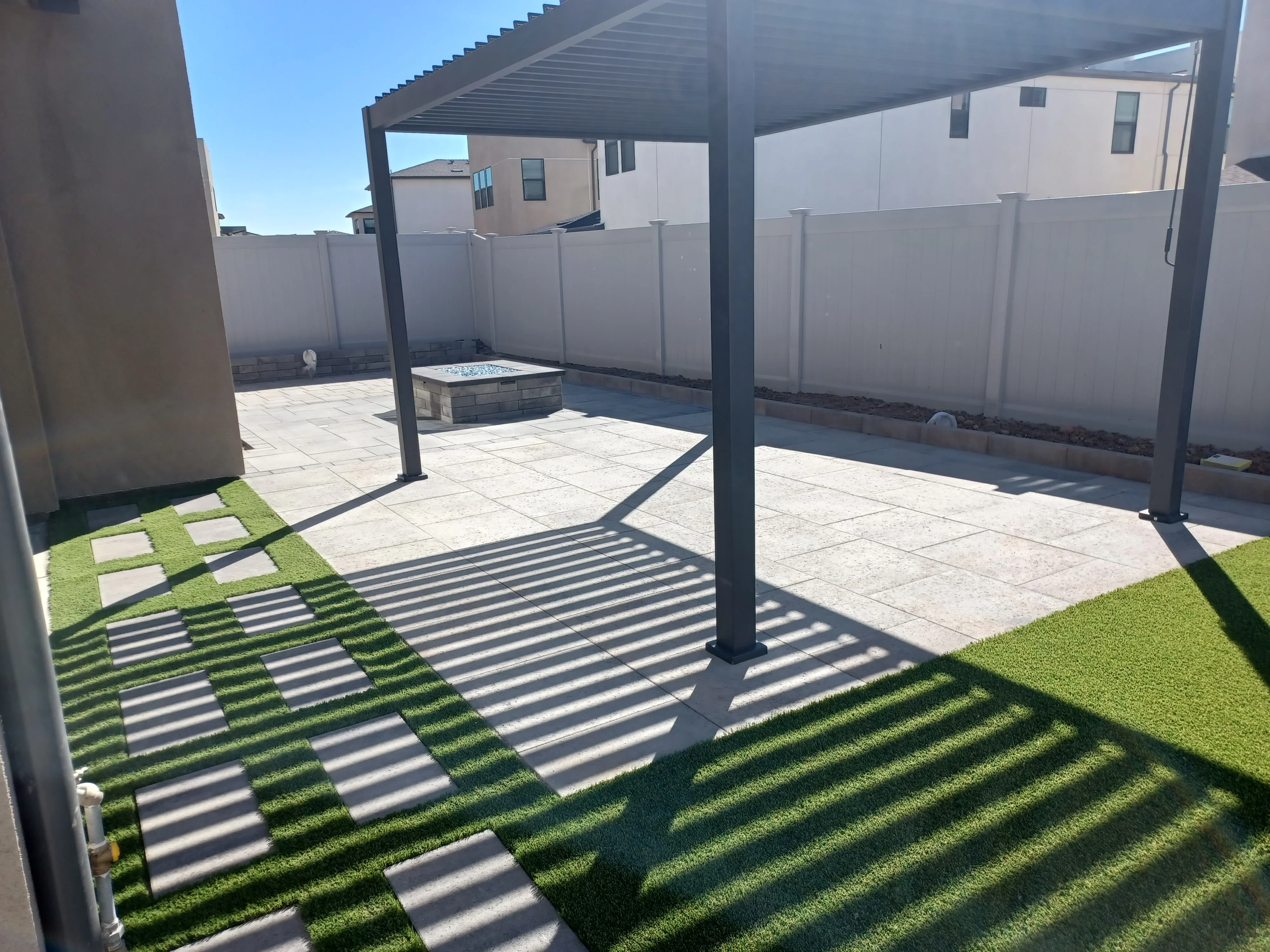 Paver patio with artificial turf strips, black pergola, and stone fire pit in fenced backyard