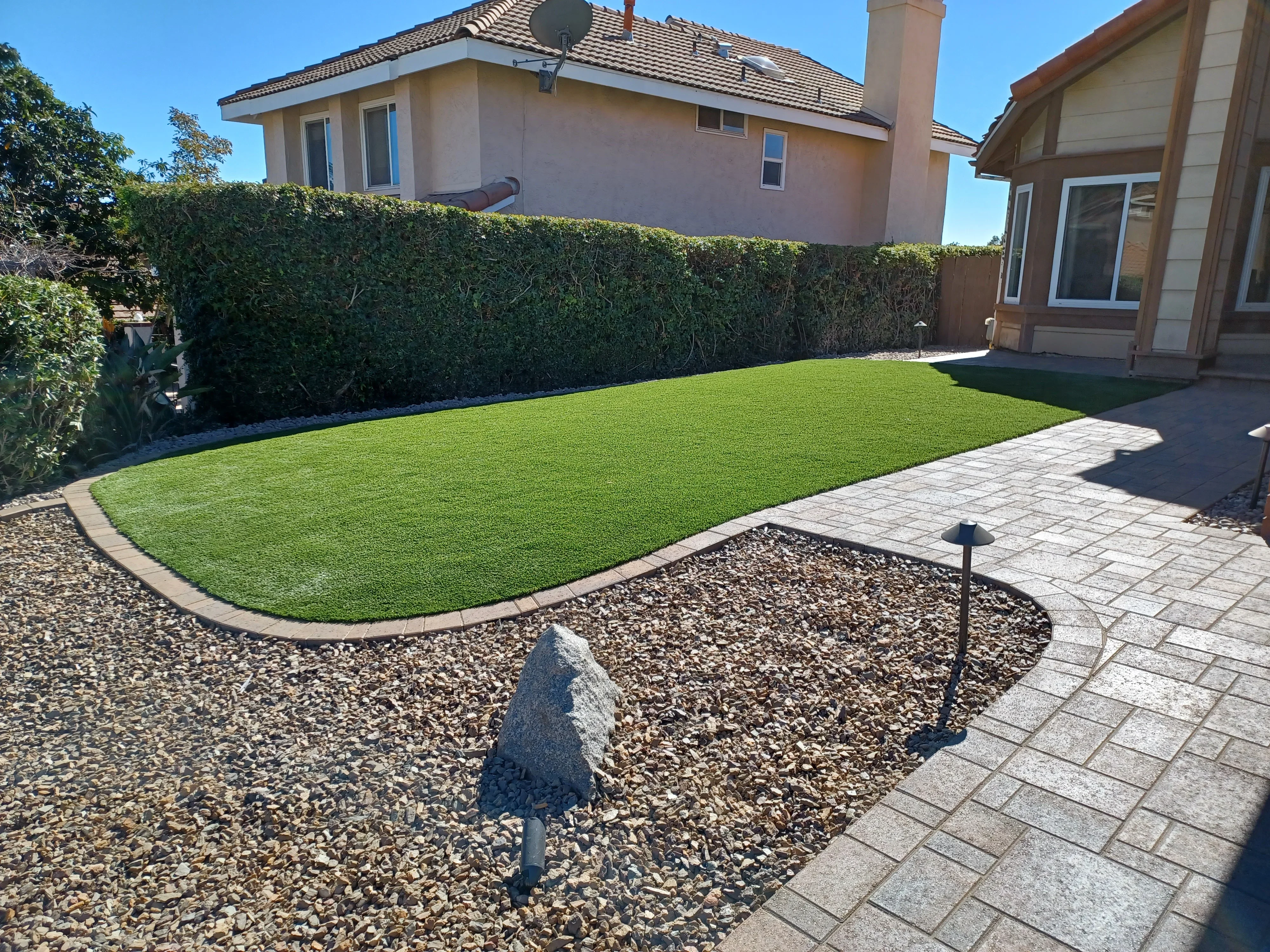 Synthetic turf lawn with concrete pavers, river rock borders, and mature hedges