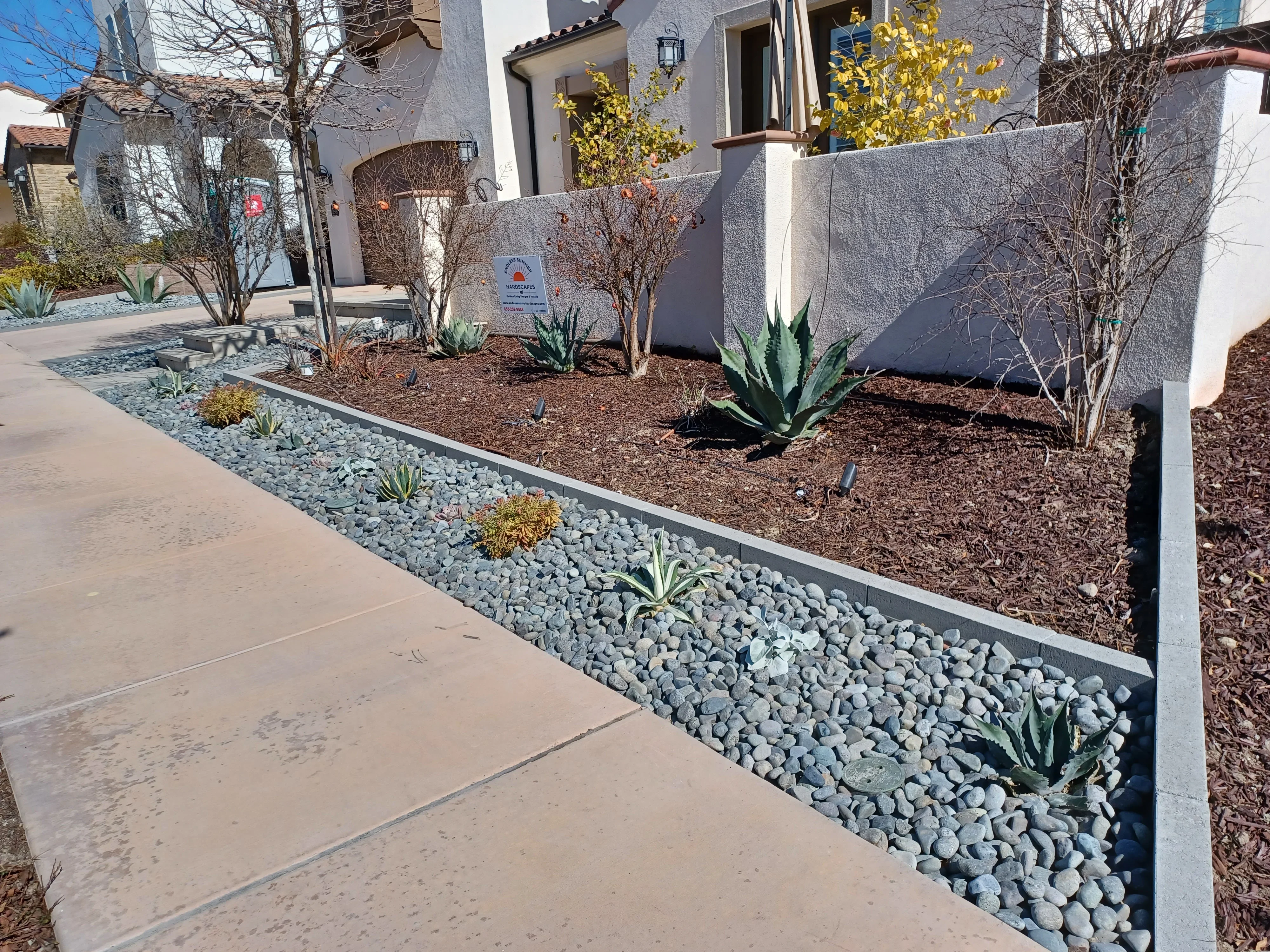 Metal-edged rock border with light gray river rock, concrete walkway, and xeriscaped planting beds