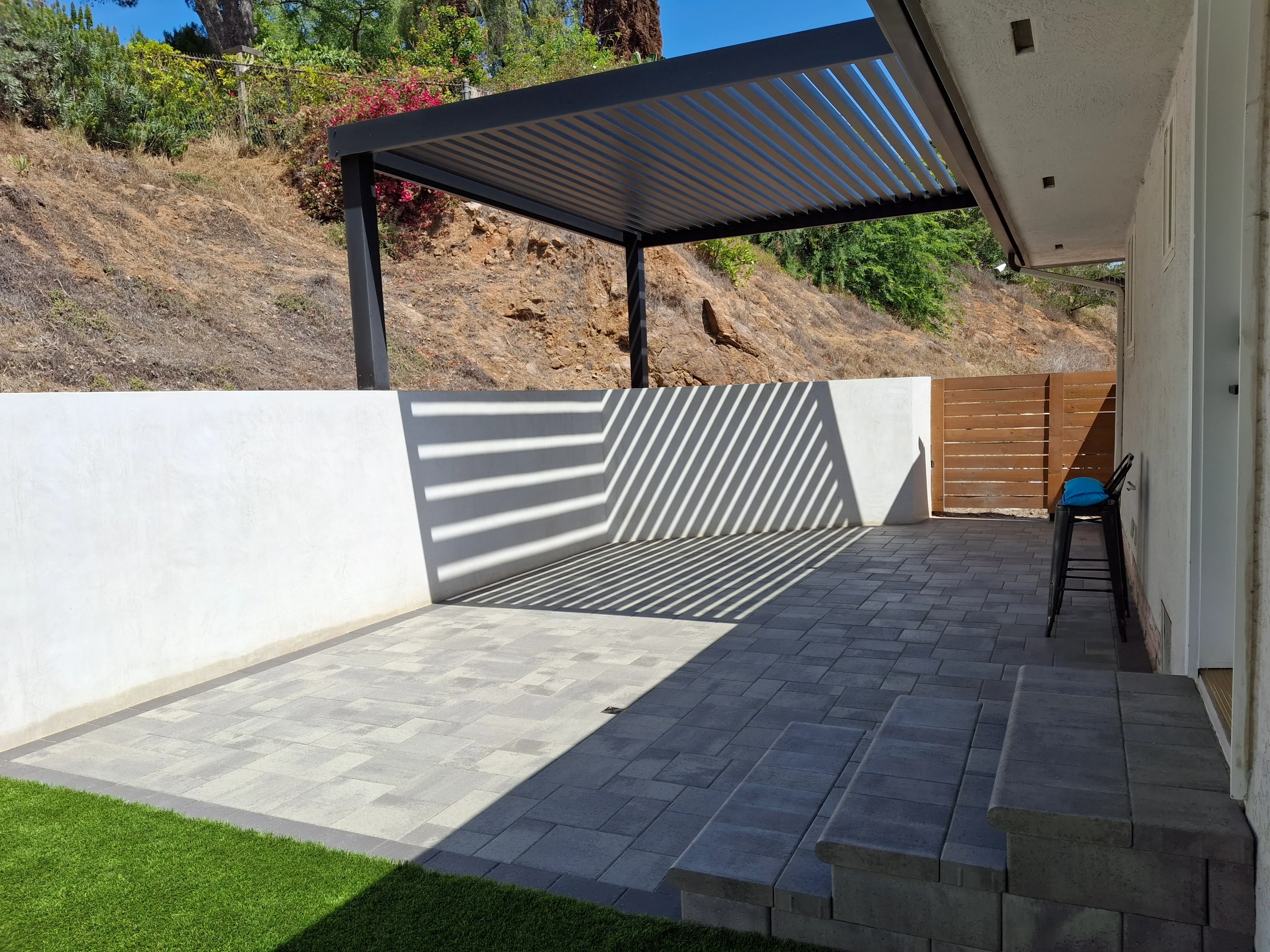 Black pergola with louvered roof over tiered paver patio, retaining wall, and wood screen