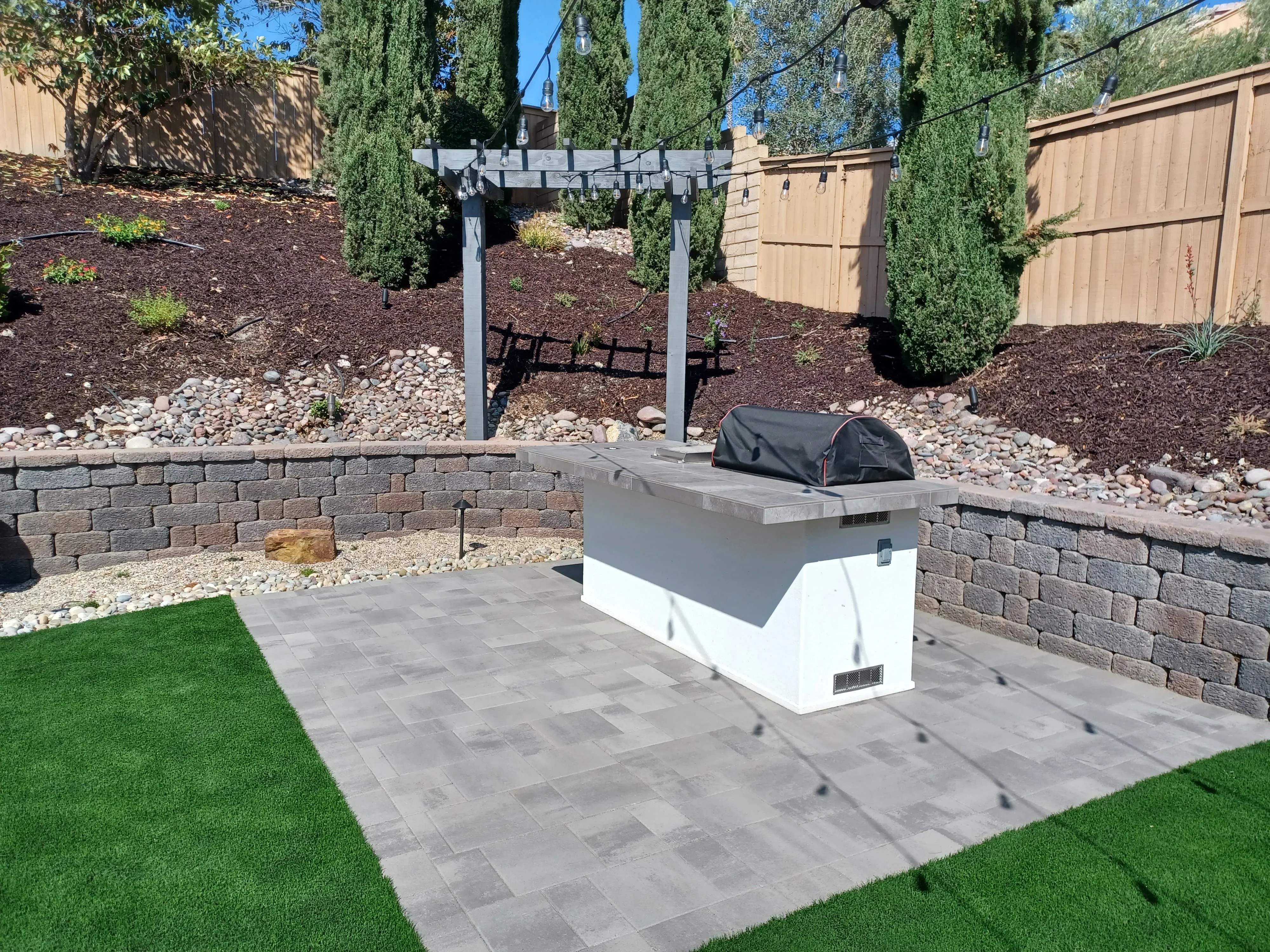 Gray paver patio with stacked stone retaining walls, built-in seating, and wooden pergola in backyard