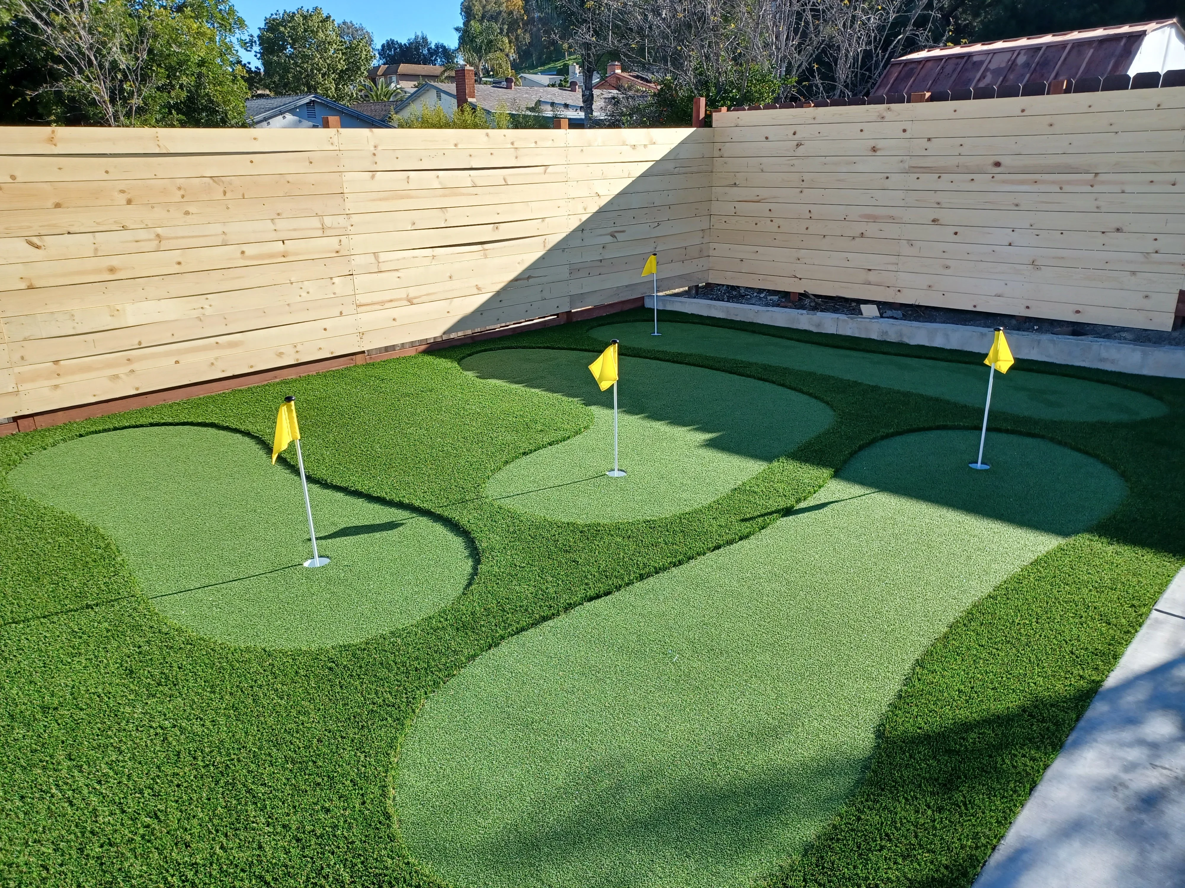 Synthetic turf putting green with contoured holes, yellow flags, timber retaining walls, and concrete edging