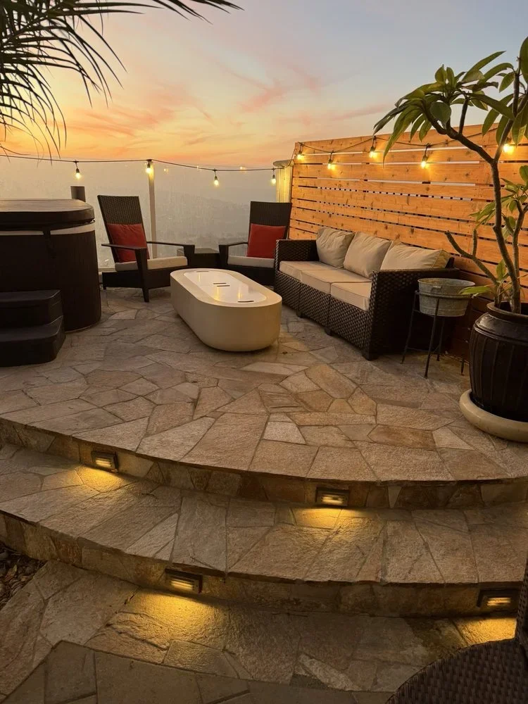 Stone patio with geometric pavers, step lighting, and wooden privacy fence at dusk
