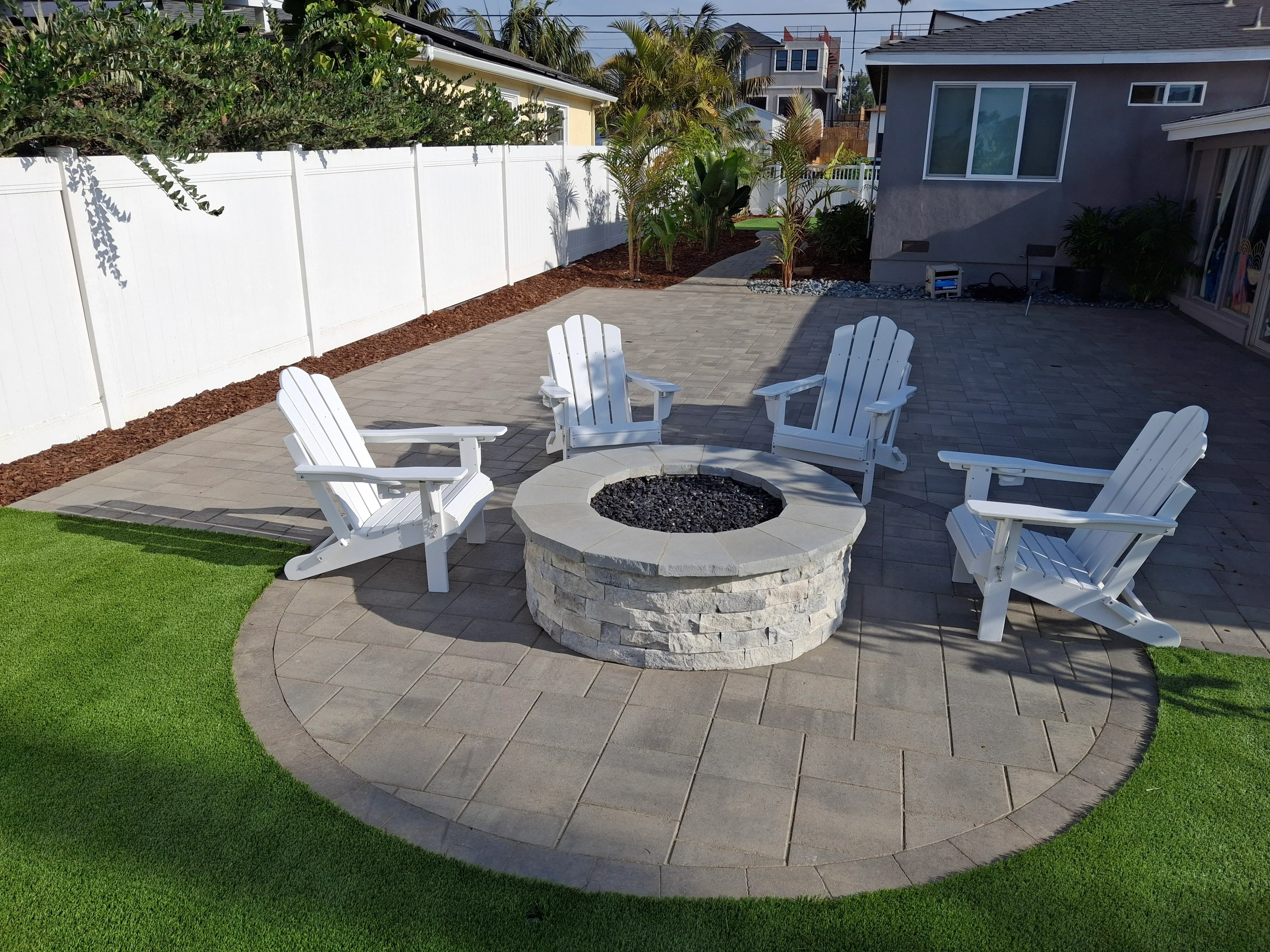 Circular paver patio with central stone fire pit, herringbone pattern pavers, and four white Adirondack chairs