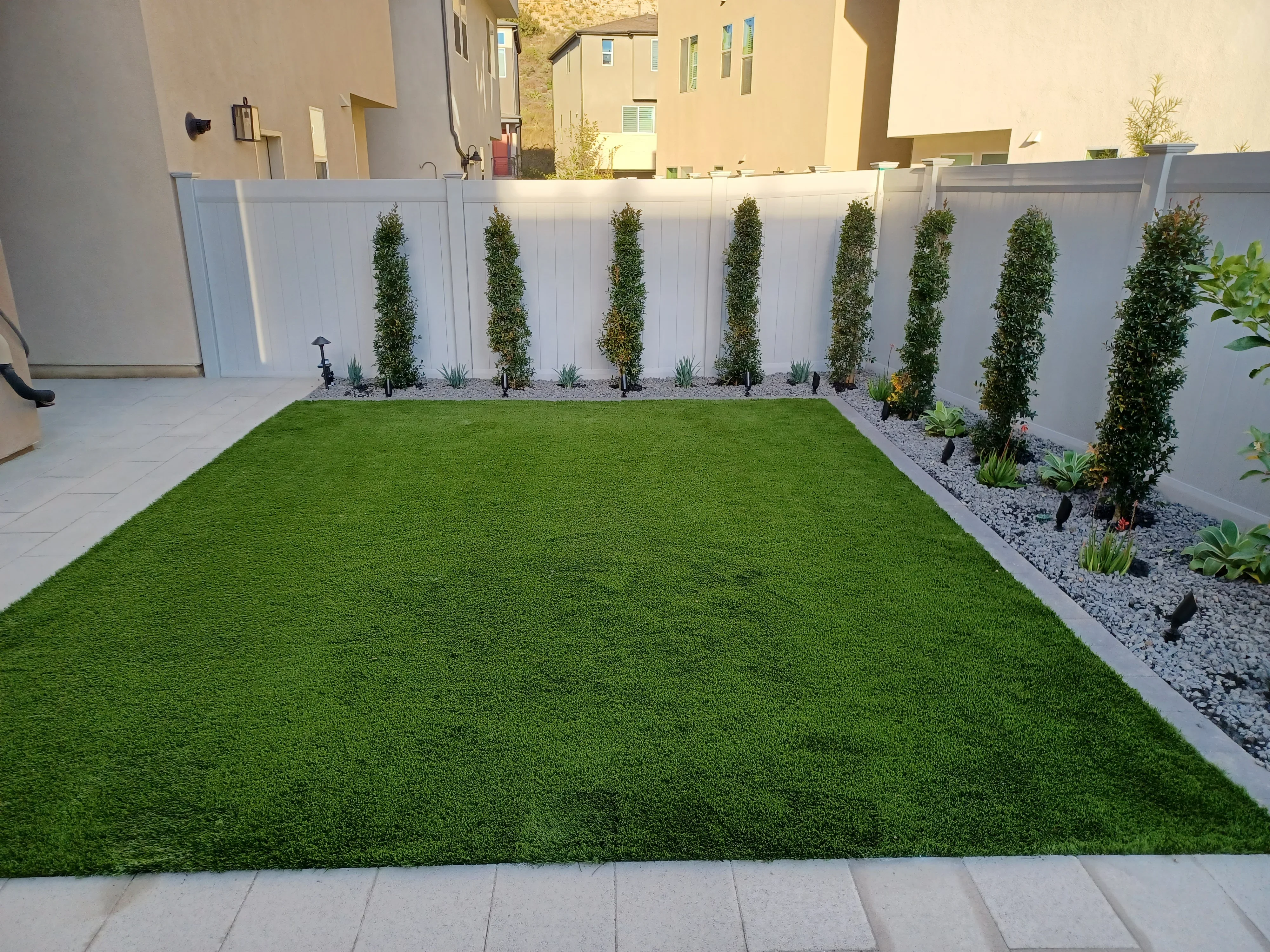 Synthetic turf lawn with white vinyl fence, columnar trees, river rock beds, and concrete paver edging