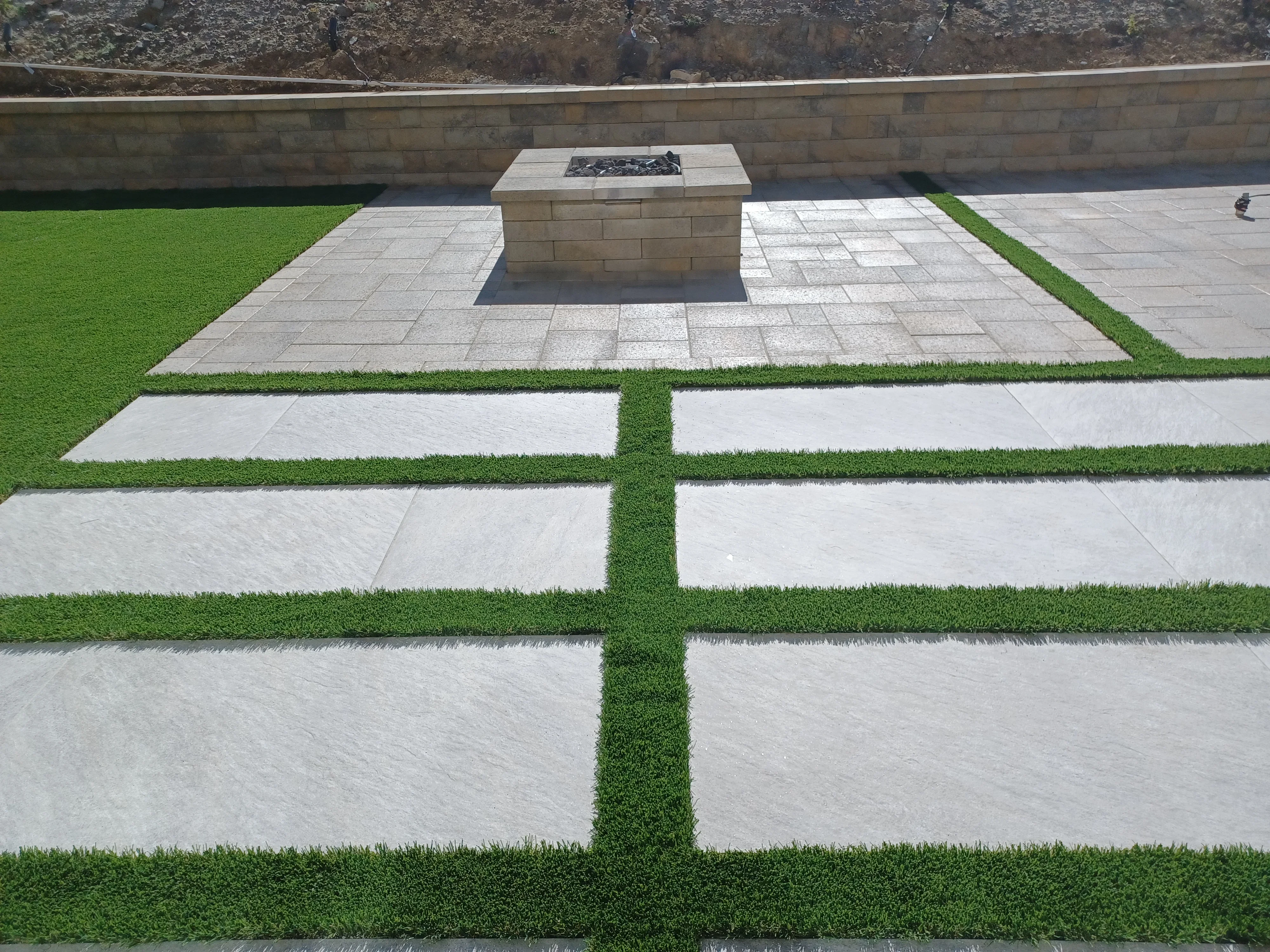 Concrete paver patio with geometric turf strips and central stone fire pit surrounded by retaining walls
