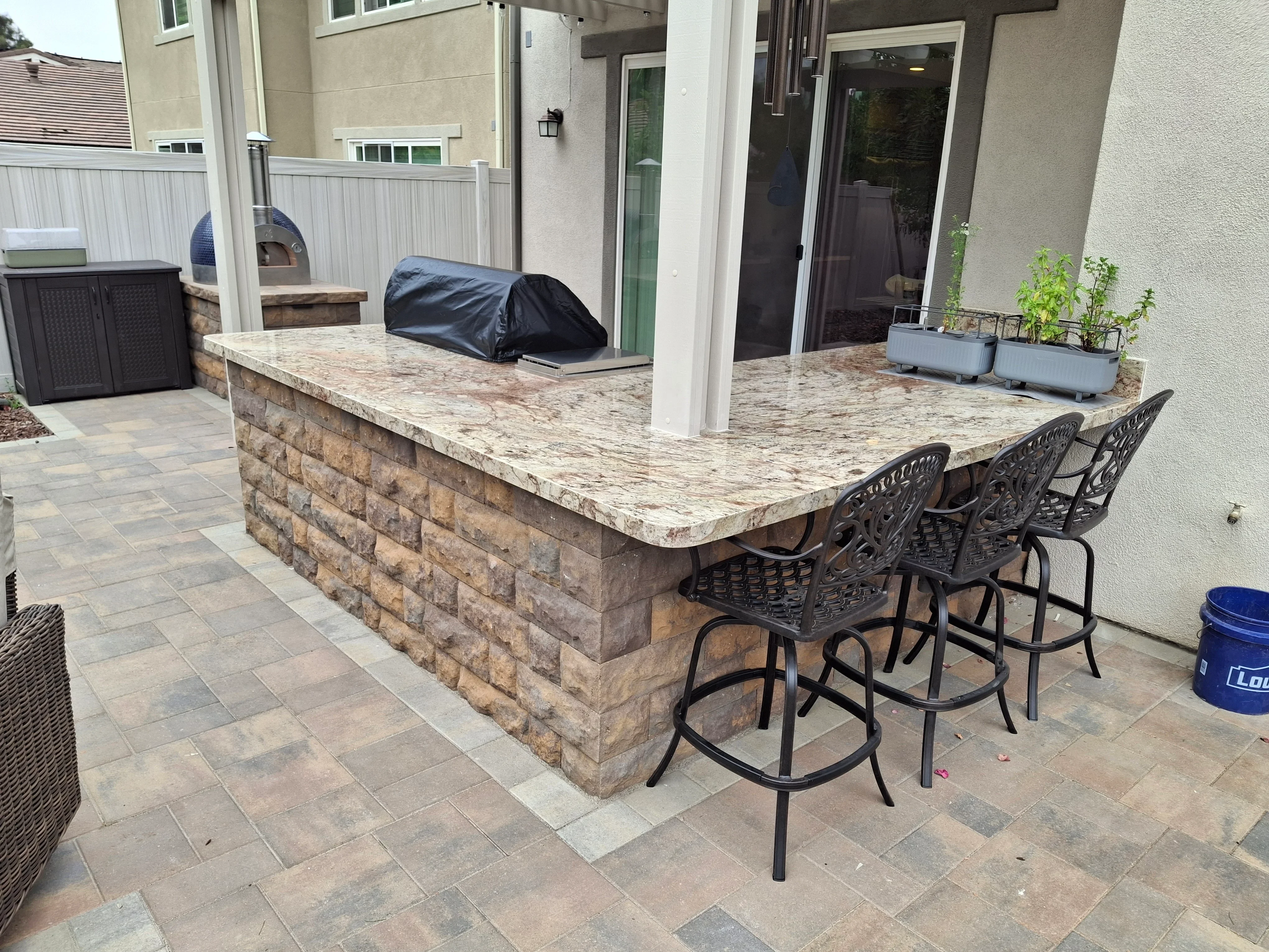 Granite-topped outdoor bar with stacked stone base and paver patio, featuring bar seating and pizza oven