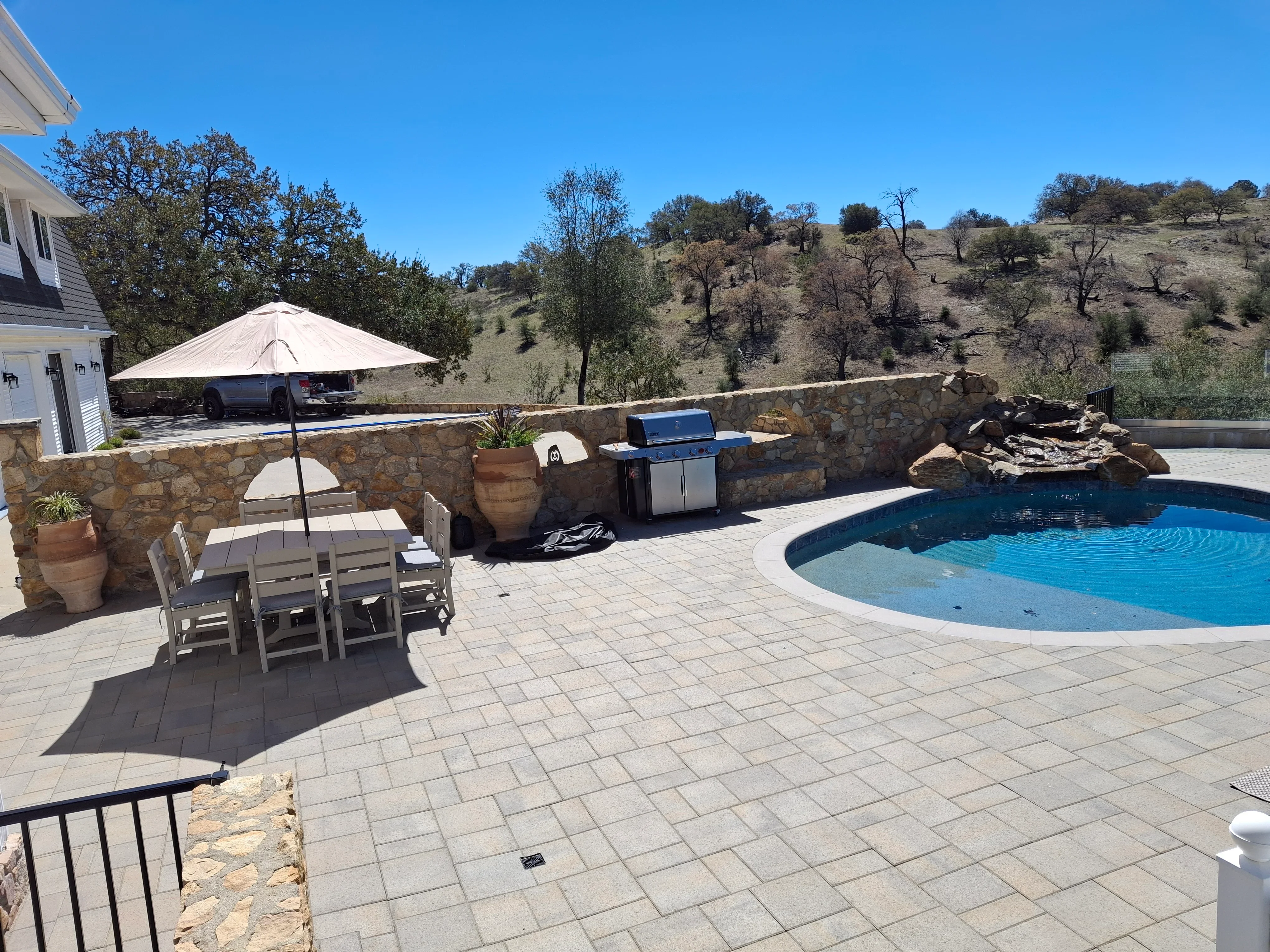 Paver pool deck with stone retaining walls, built-in grill, and curved pool on hillside