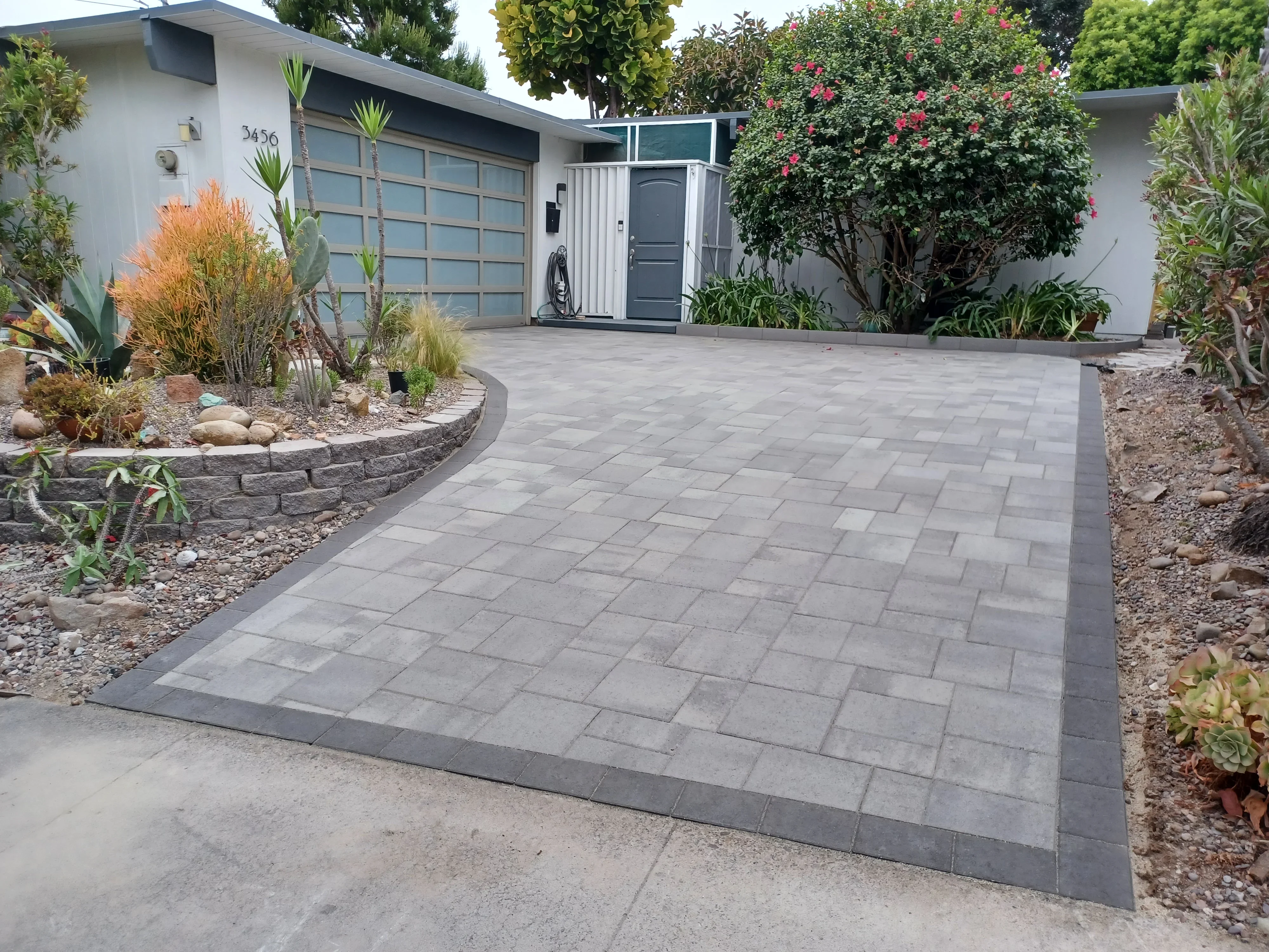 Gray concrete paver driveway with dark border, xeriscape plantings, and stone retaining walls