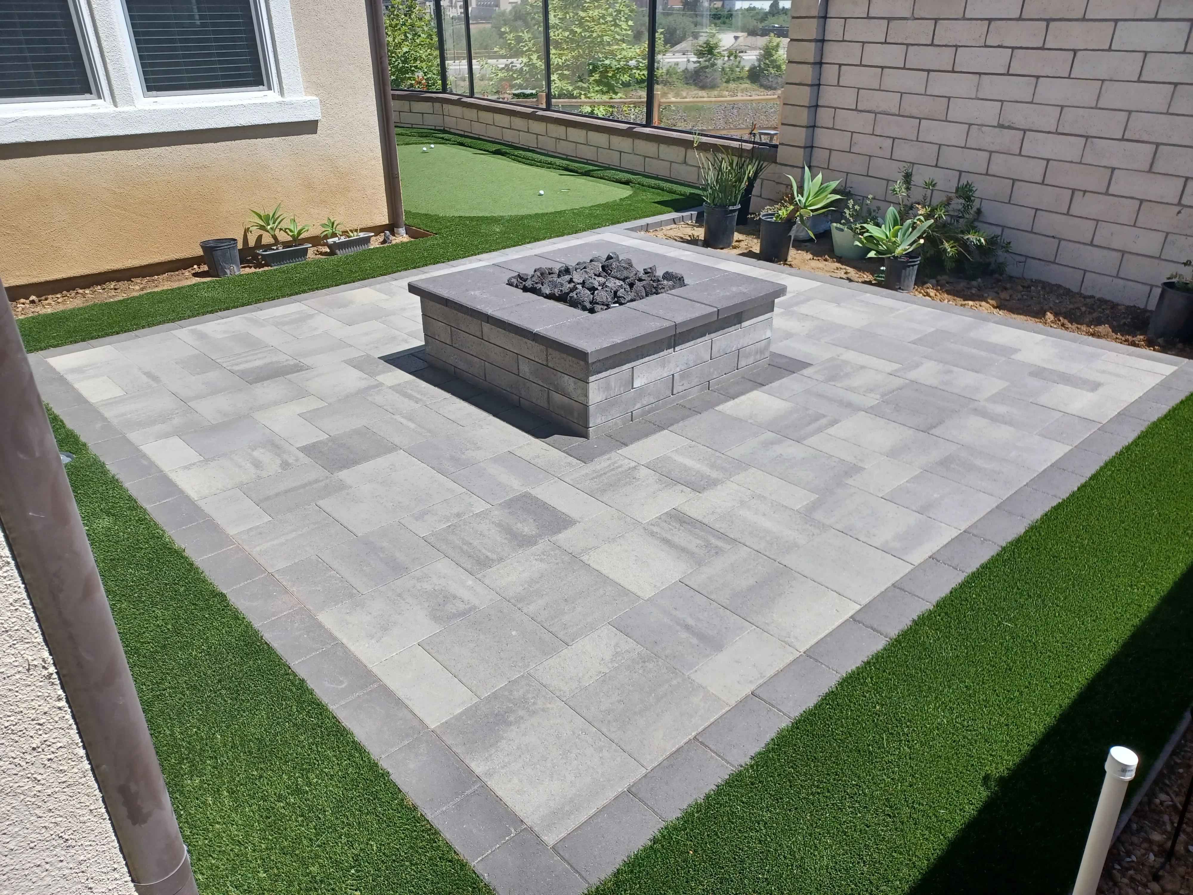 Gray concrete paver patio with central fire pit, bordered by synthetic grass and potted plants