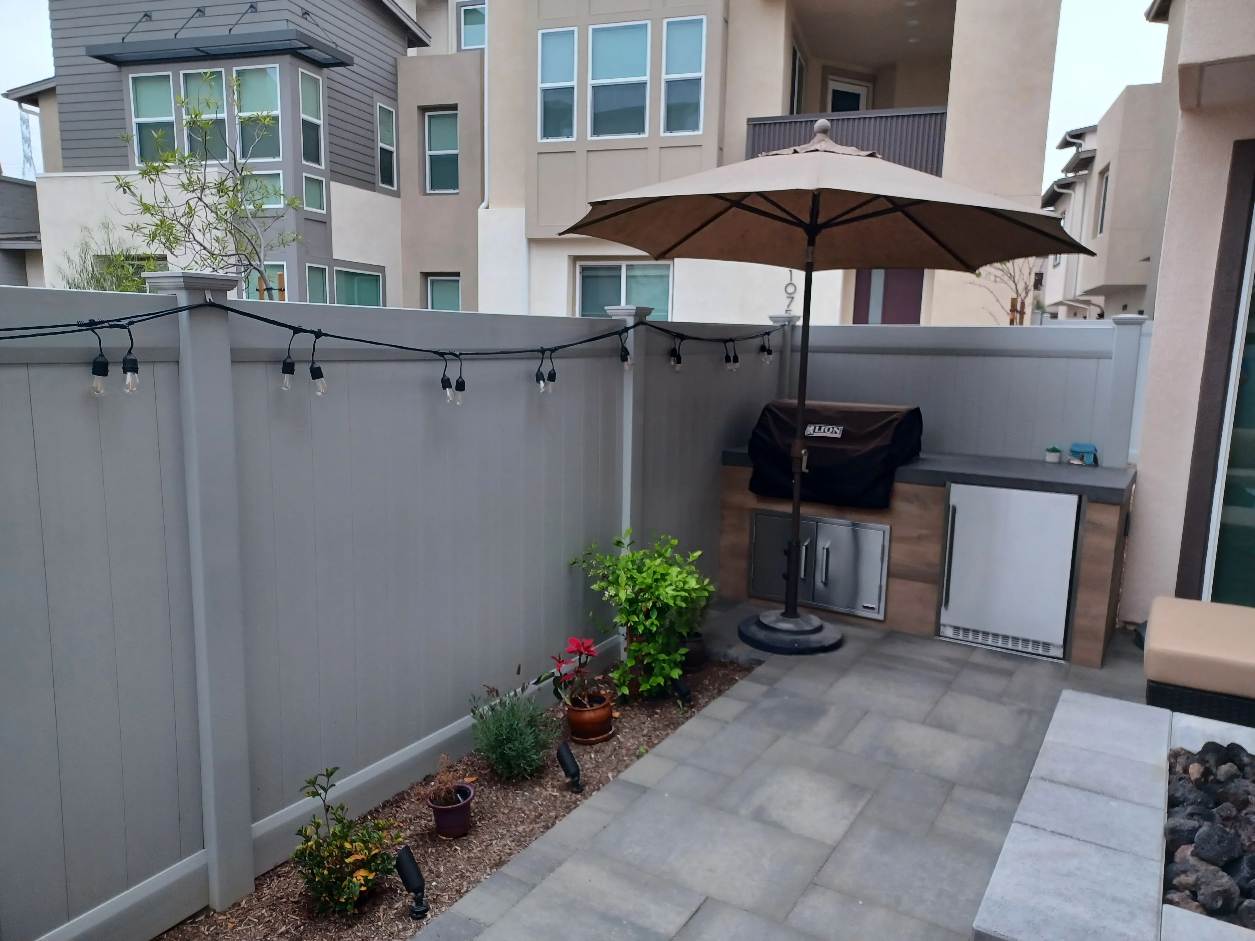 Gray paver patio with vinyl privacy fence, string lights, outdoor kitchen with stainless steel grill and refrigerator