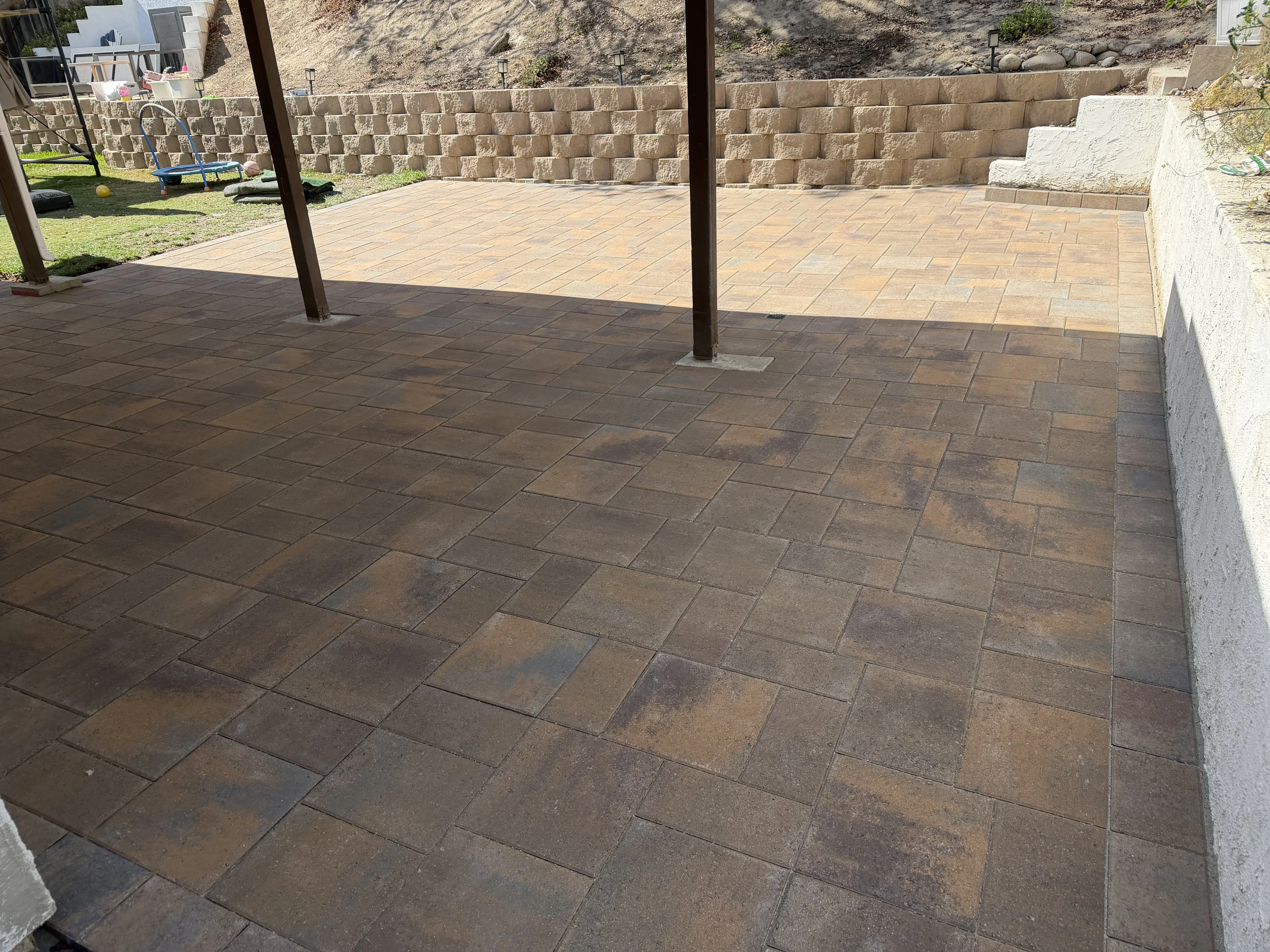 Dark brown paver patio with running bond pattern, wooden posts, and tan stone retaining walls on hillside