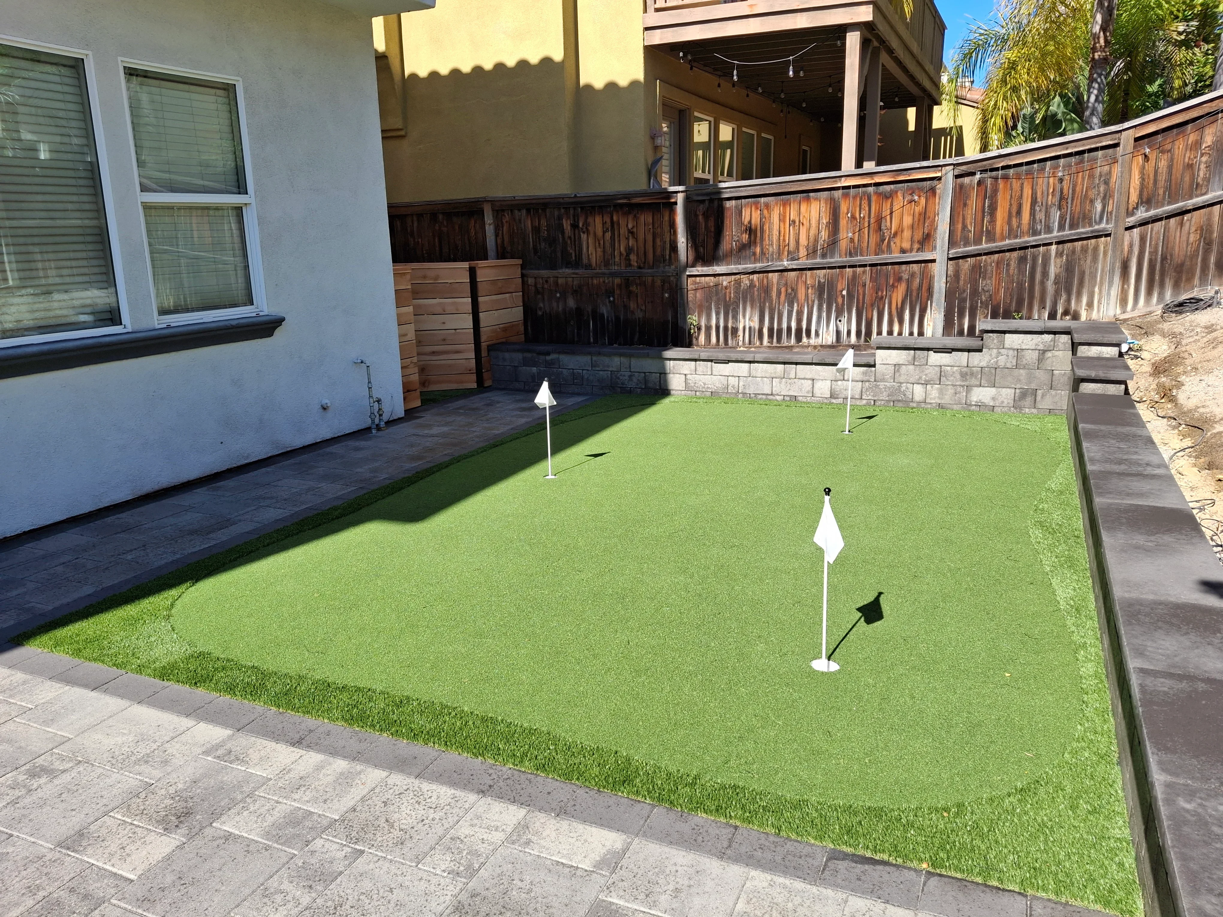 Synthetic putting green with concrete paver borders and stone retaining wall in residential backyard