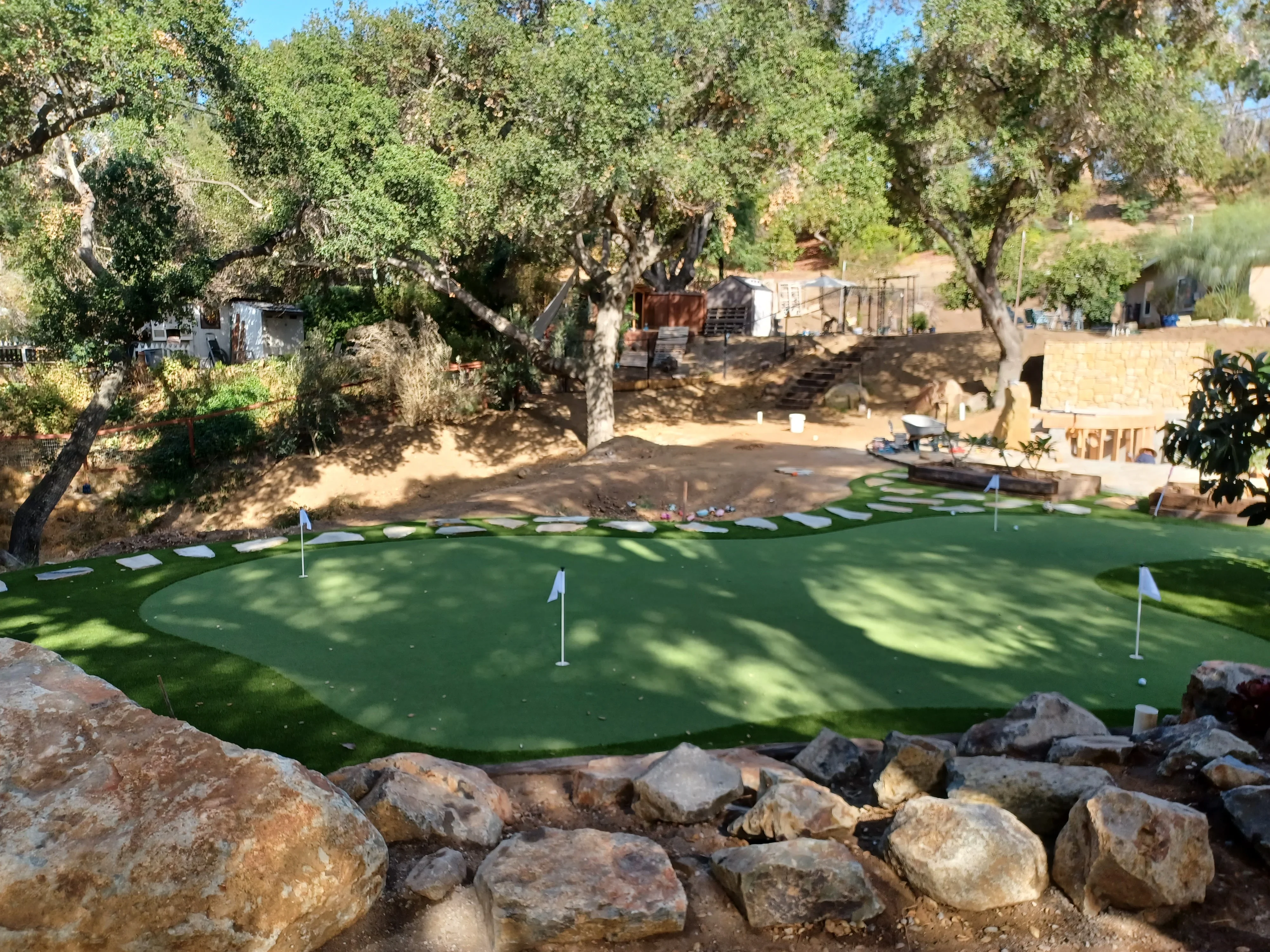 Synthetic putting green with flagged holes, natural boulder border, and mature oak trees