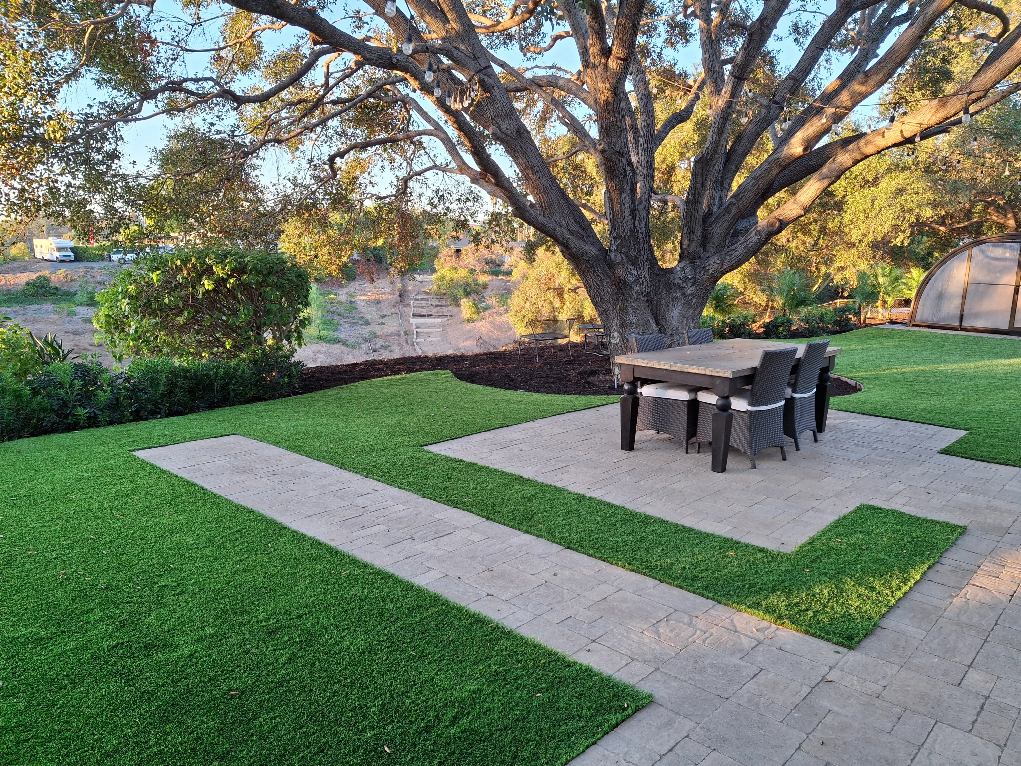 Gray pavers in geometric pattern with synthetic turf, mature oak tree, outdoor dining setup