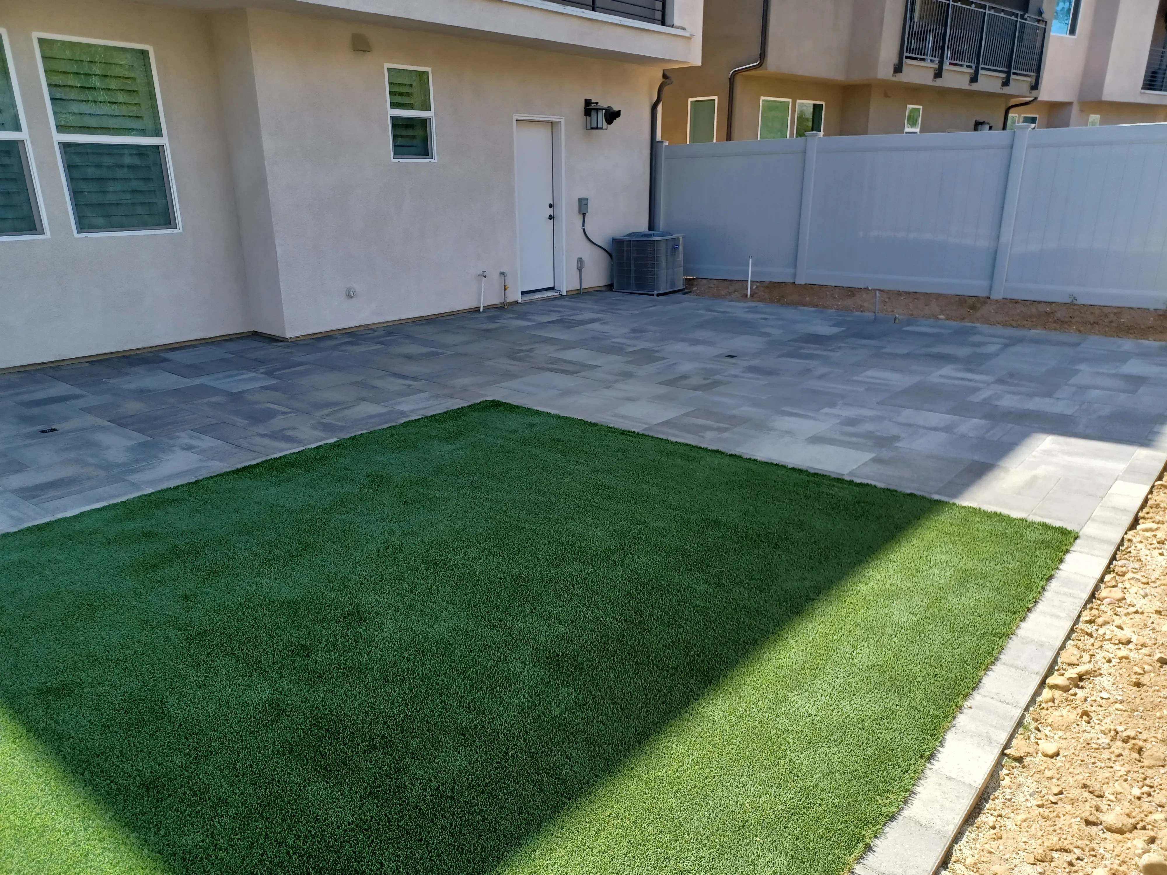 Gray composite paver patio with synthetic turf sections and concrete edging in residential backyard