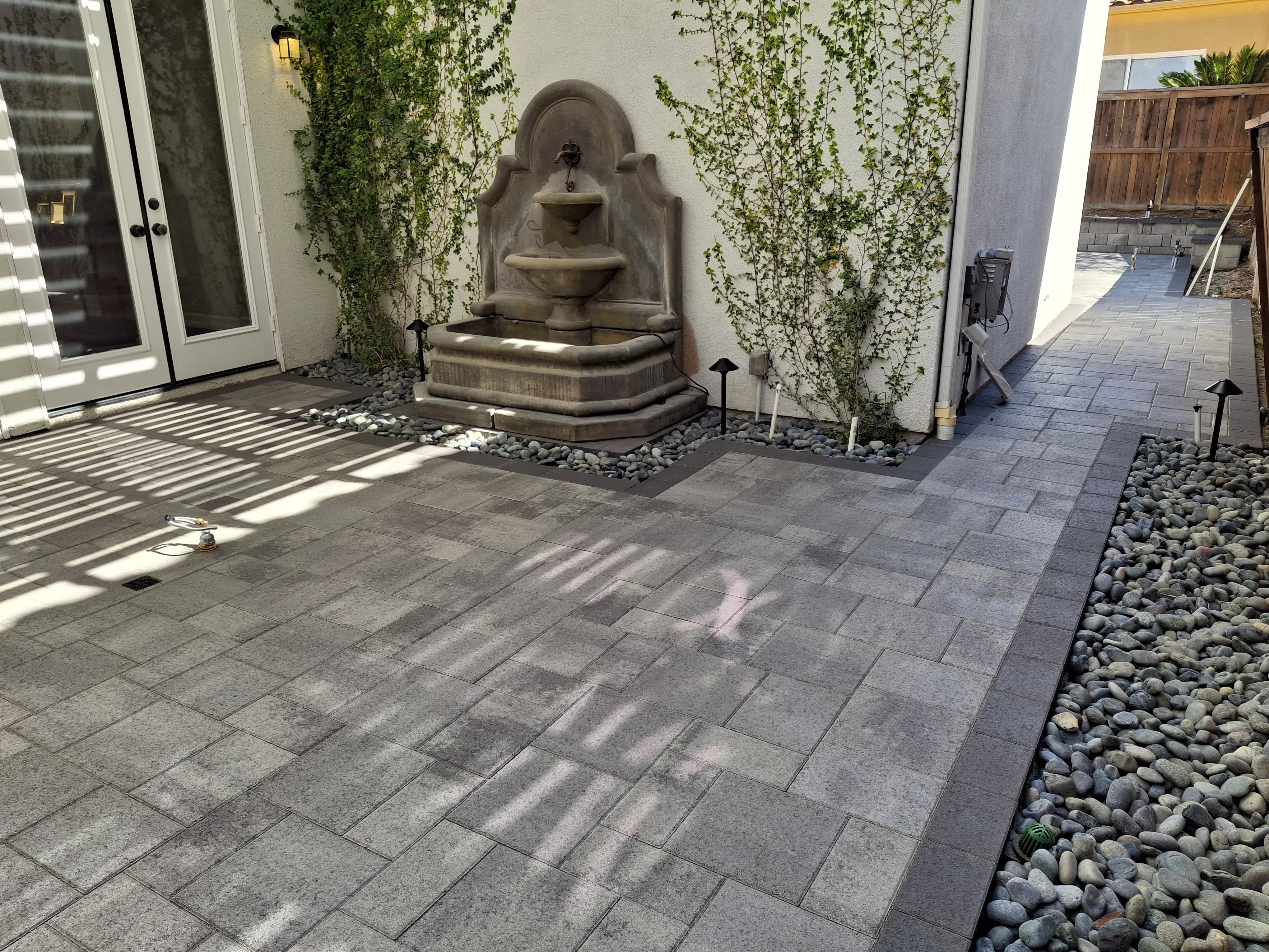 Gray paver patio with tiered stone fountain, climbing ivy, river rock, and landscape lighting