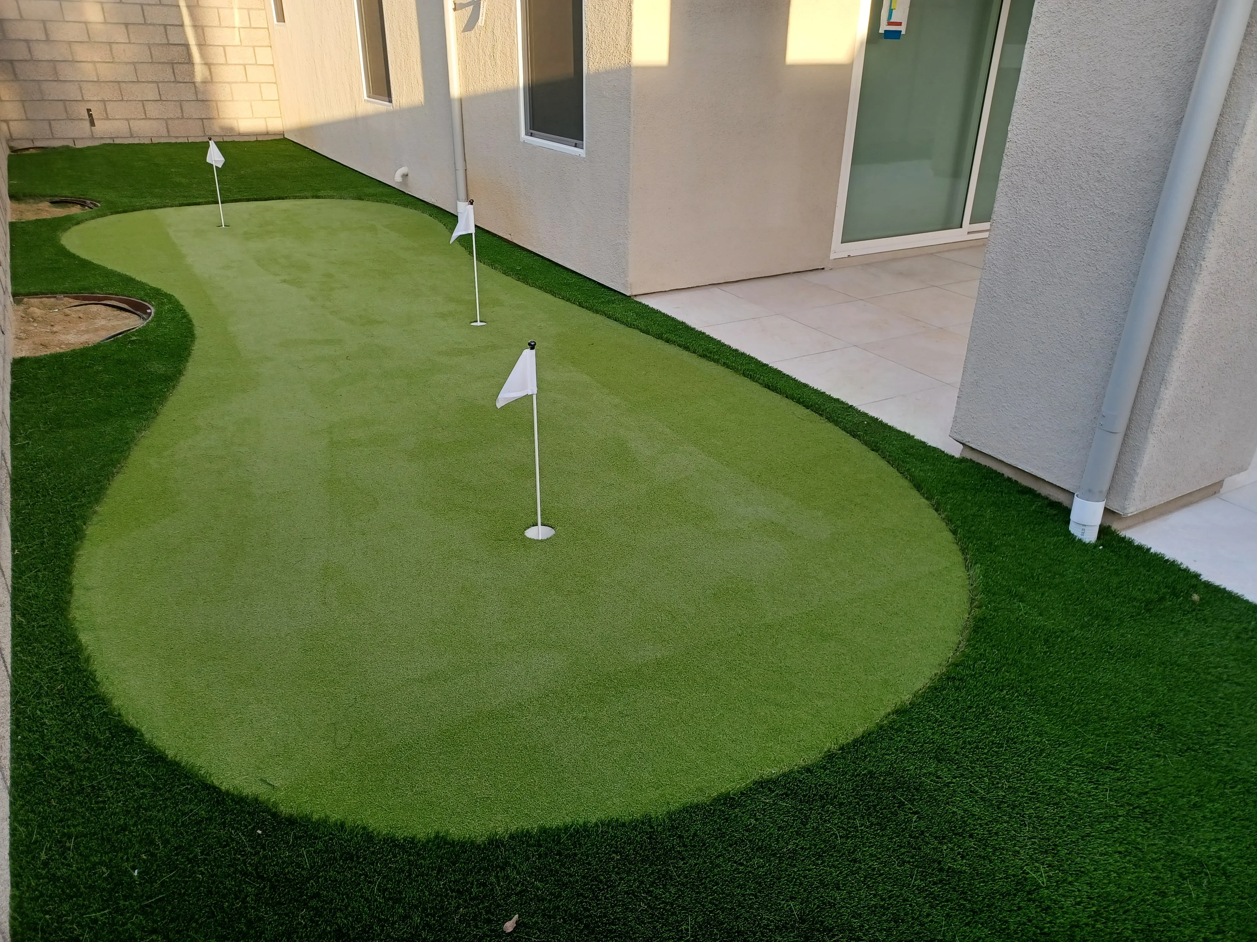 Synthetic putting green with two flagged holes, sand bunker, and dark turf border adjacent to patio