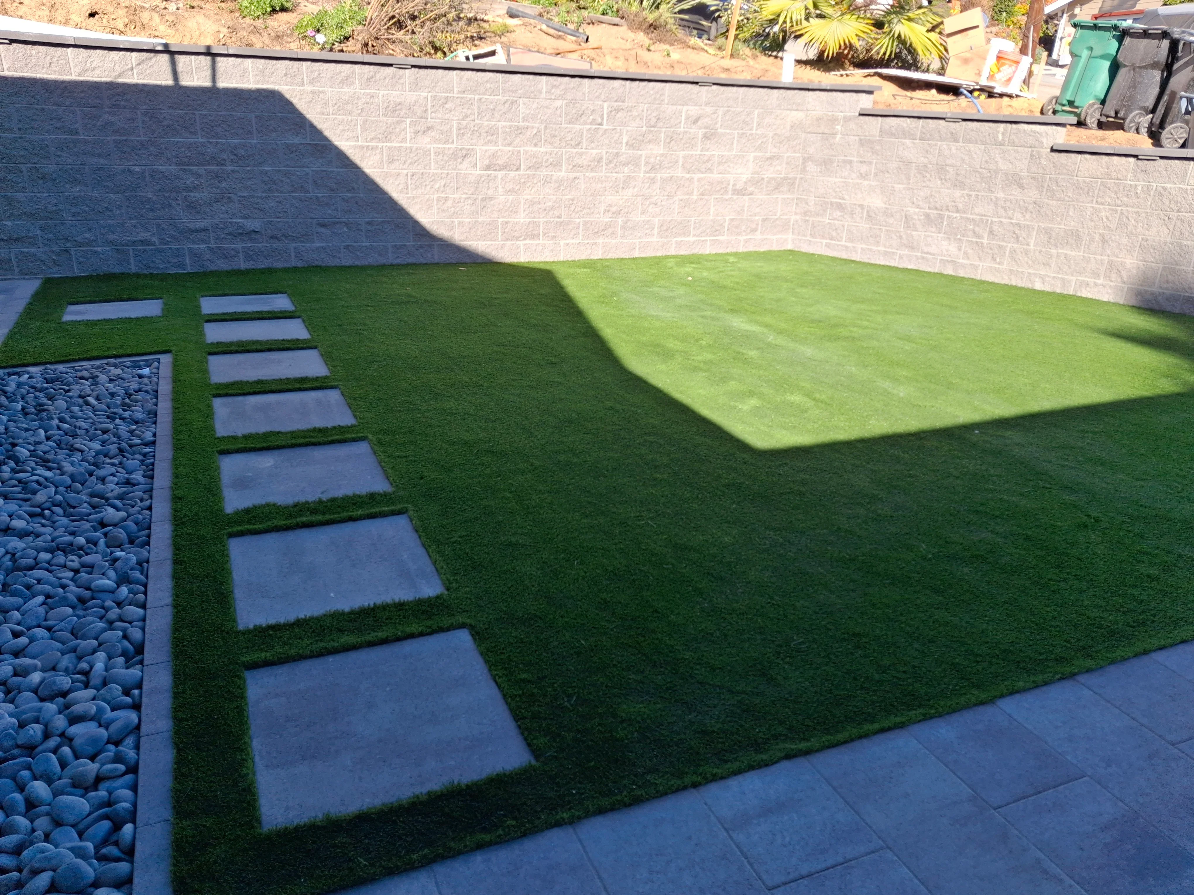 Synthetic turf yard with blue-gray stepping stone pathway, river rock bed, and concrete retaining wall