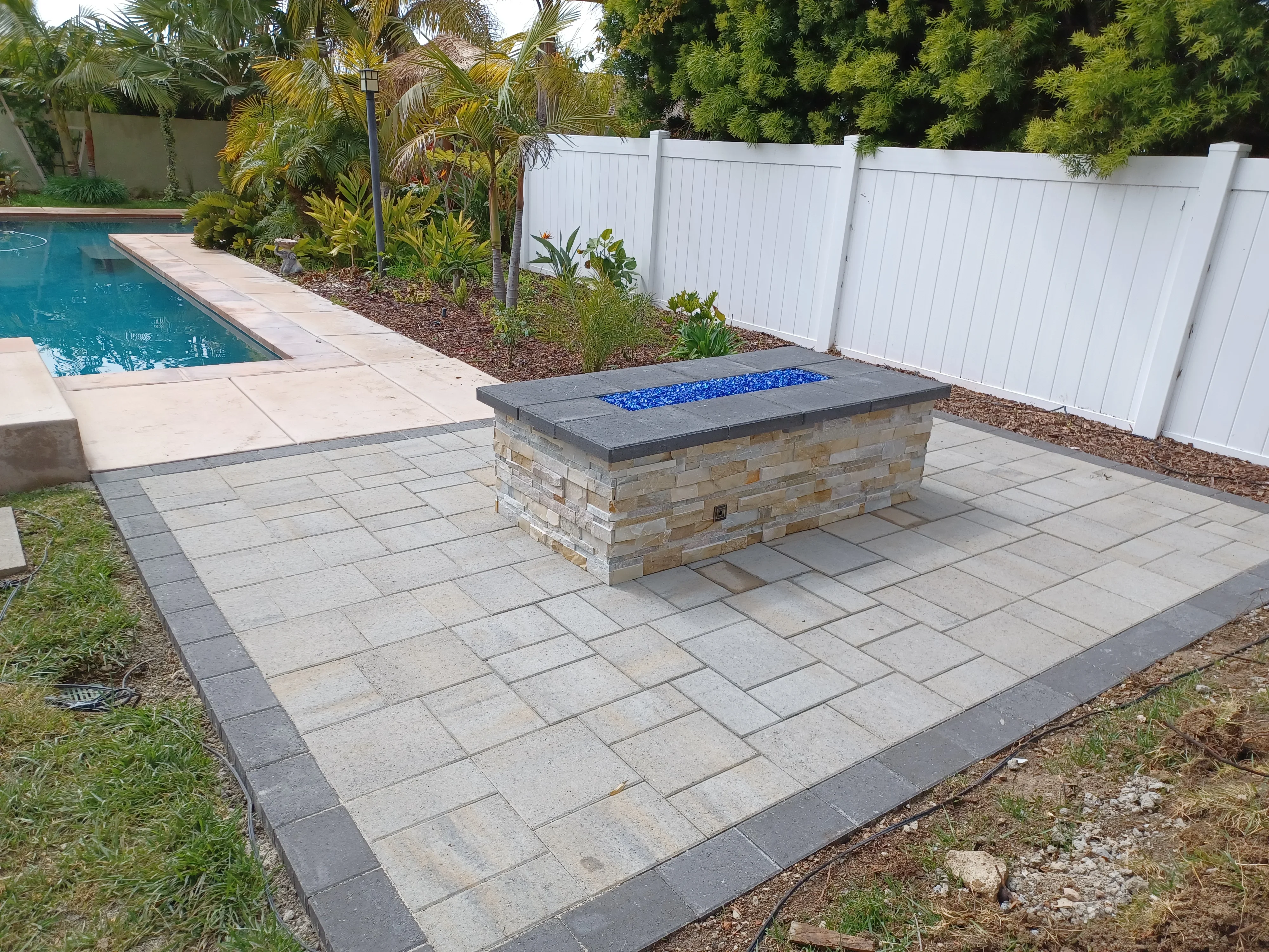 Concrete pavers with dark border and stacked stone fire pit with blue glass, poolside patio