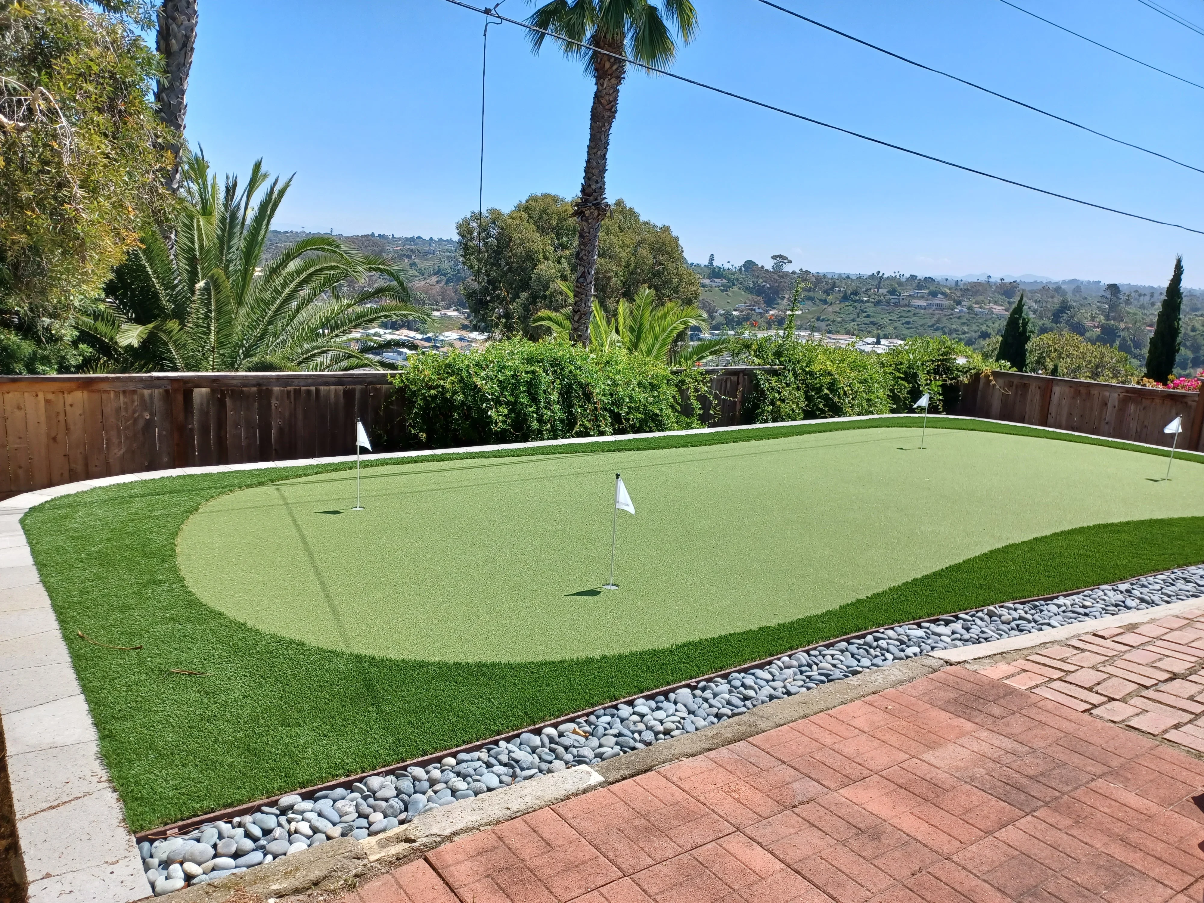 Synthetic putting green with multiple holes, river rock border, concrete patio, and wood fence